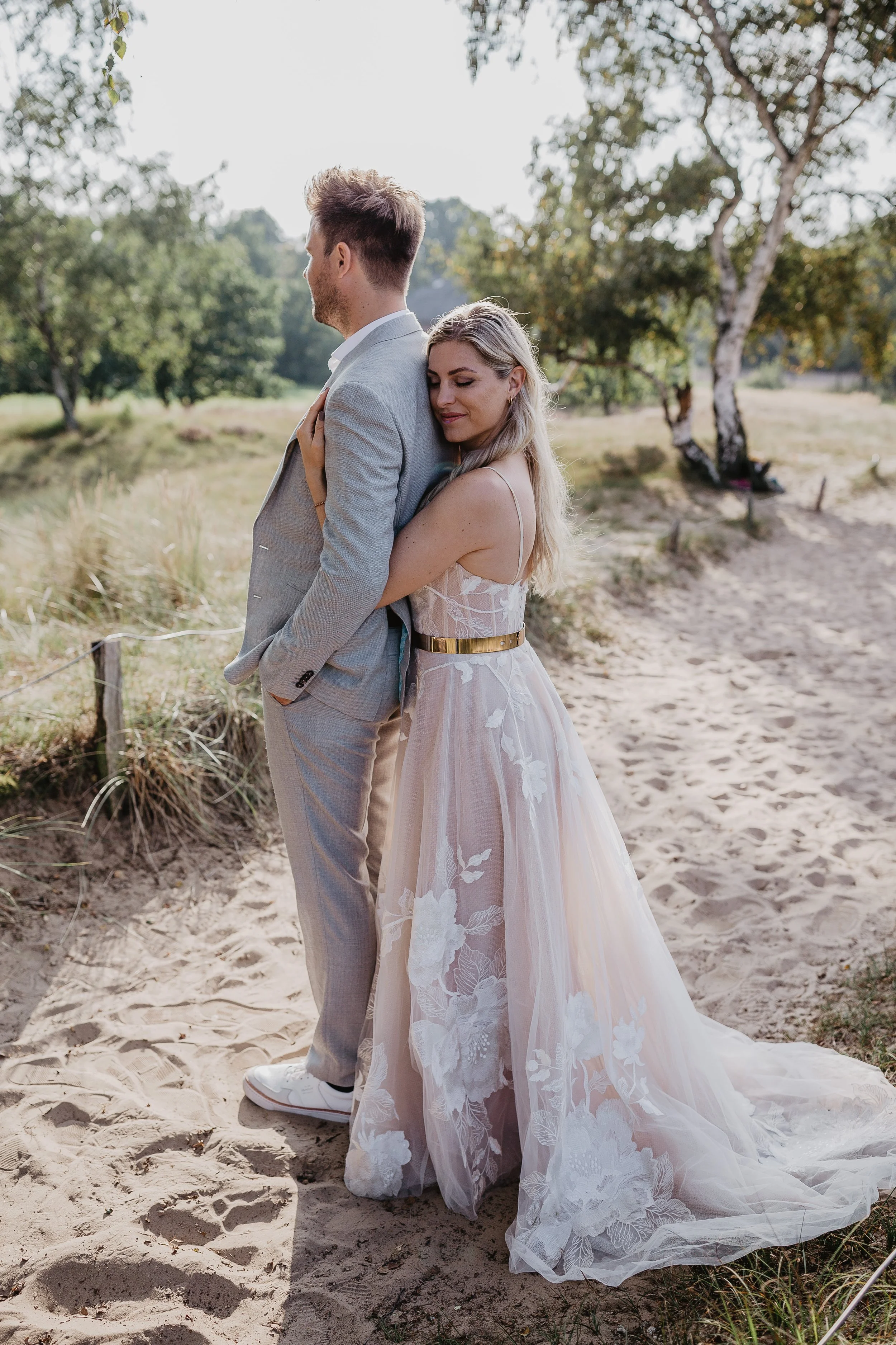 Ein Paar in formeller Kleidung steht im Sand in einer natürlichen Umgebung mit Bäumen, die Frau umarmt den Mann von hinten, das Bild vermittelt eine intime und romantische Stimmung.
