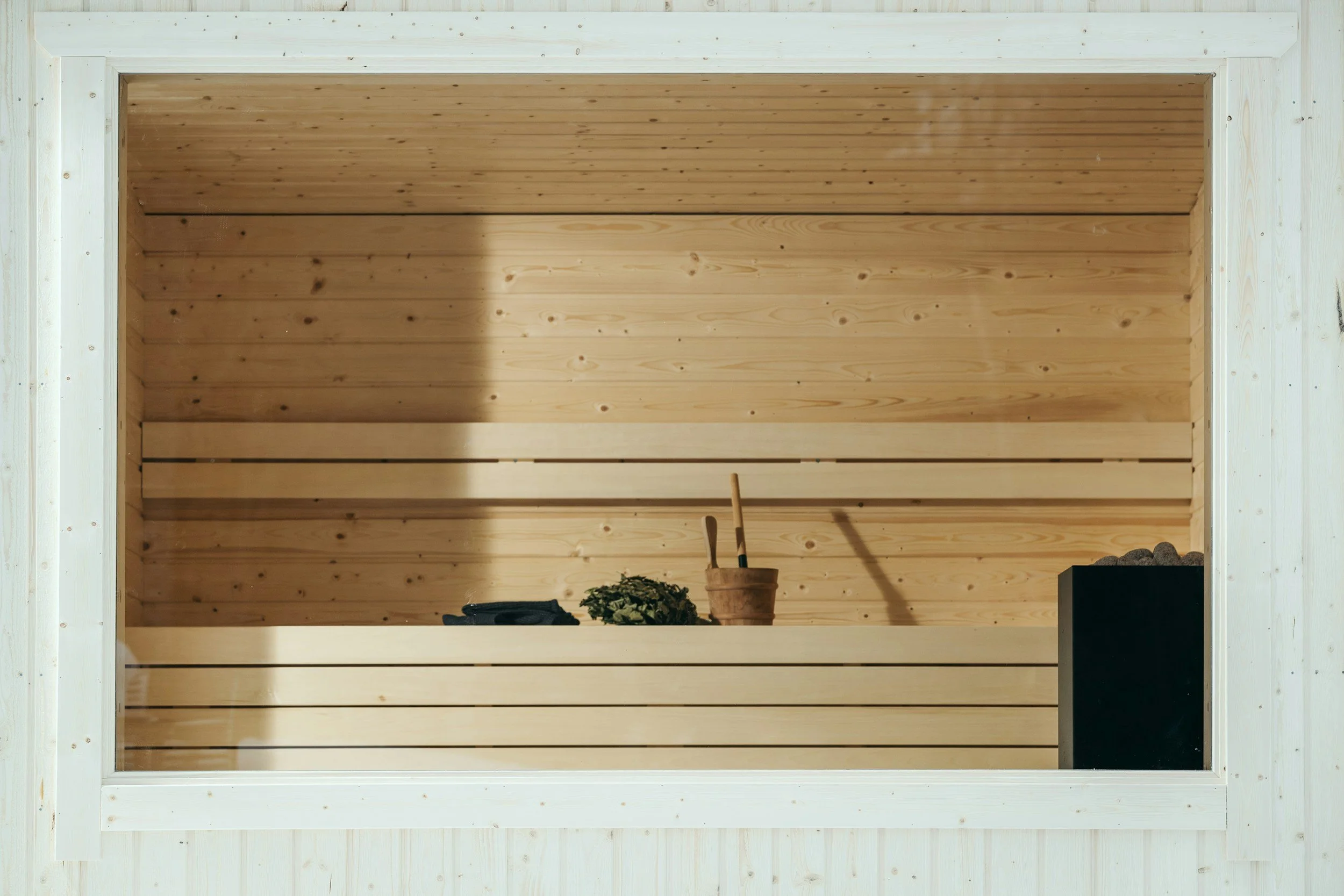 Interior of a wooden sauna with a bench, a small plant, a towel, a bucket with sauna accessories, and a heater with stones.
