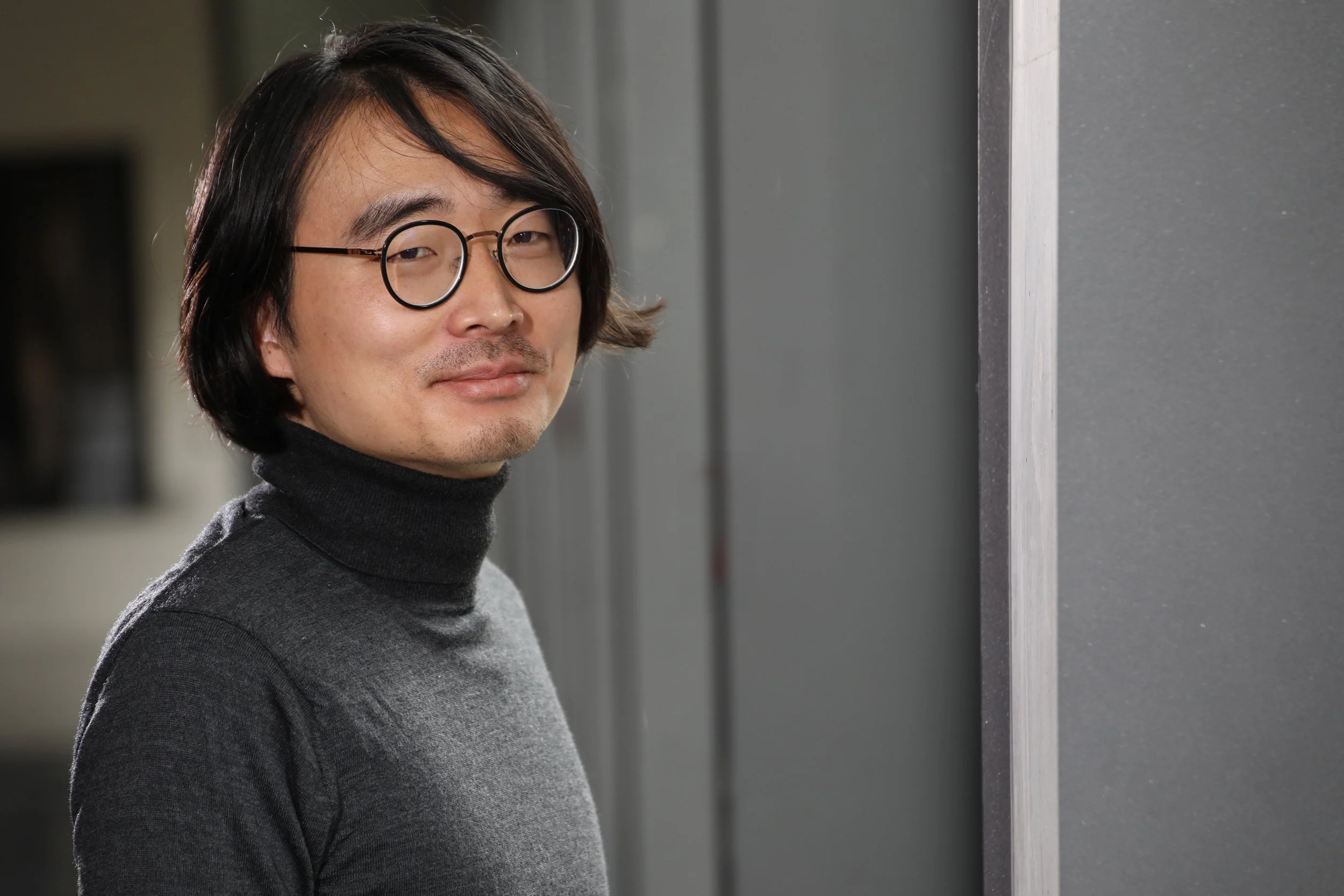 Ein Mann mit schwarzen, mittellangen Haaren, Brille und dunklem Rollkragenpullover steht in einer modernen Umgebung.