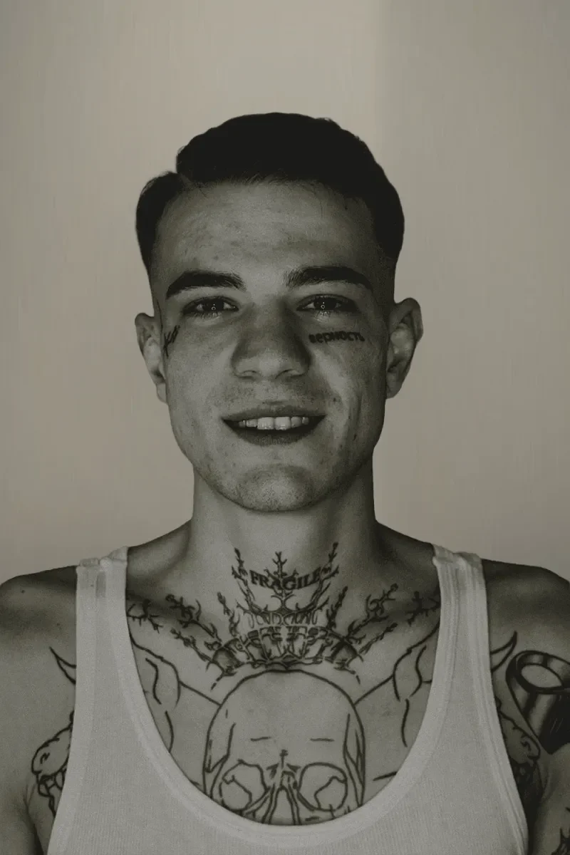 Schwarz-Weiß-Fotografie eines jungen Mannes mit Tätowierungen im Oberkörper, der weißes Tanktop trägt, lächelt und direkt in die Kamera schaut.