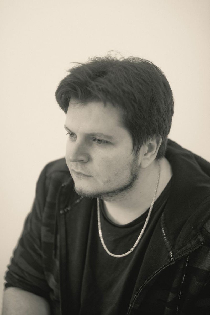 Schwarzer und weißer Portrait eines jungen Mannes mit dunklem Haar, Bart, in dunkler Kleidung und mit Halskette, blickt nach links.