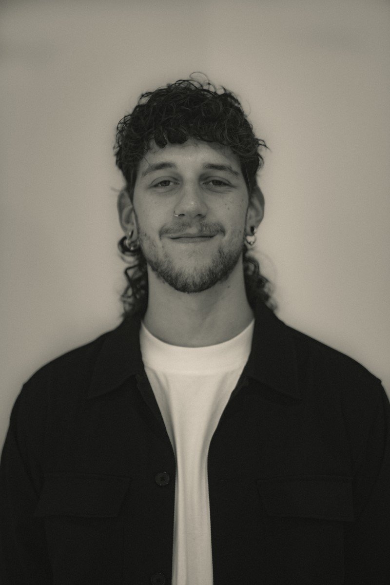 Schwarz-weiß Foto eines lächelnden jungen Mannes mit trautem Blick, dunklen Locken, Ohrpiercings, Nase und Kinnbart, trägt eine schwarze Jacke und ein weißes T-Shirt, vor neutralem Hintergrund.