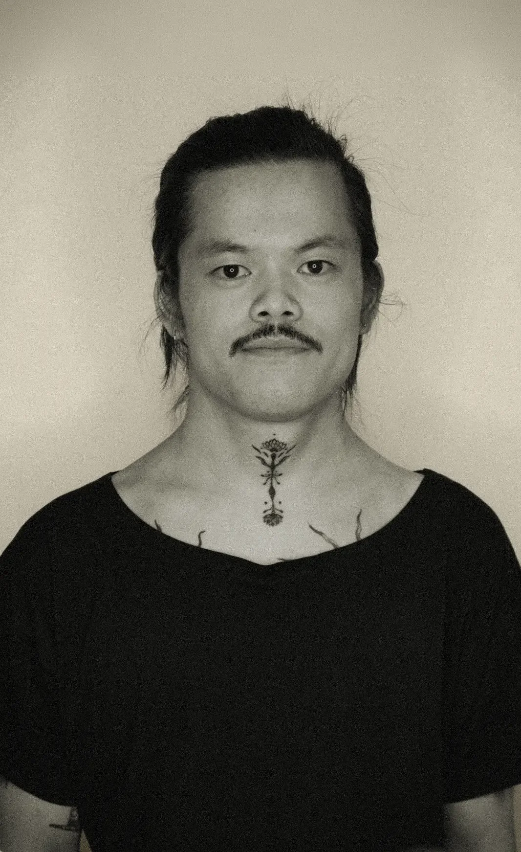 Schwarz-weiß-Foto eines Mannes mit schwarzen Haaren, Mustache, im schwarzen T-Shirt, mit Tattoo am Hals, vor einem weißen Hintergrund.