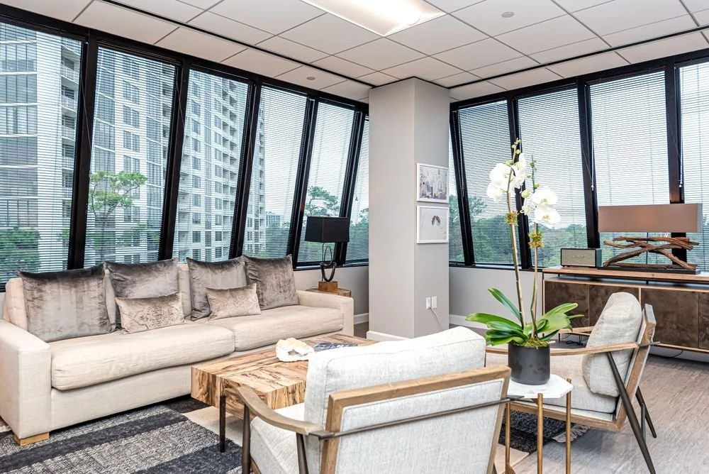 Modern living room with large angled windows, beige sofa with gray pillows, wooden coffee table, white orchid plant, and cityscape view