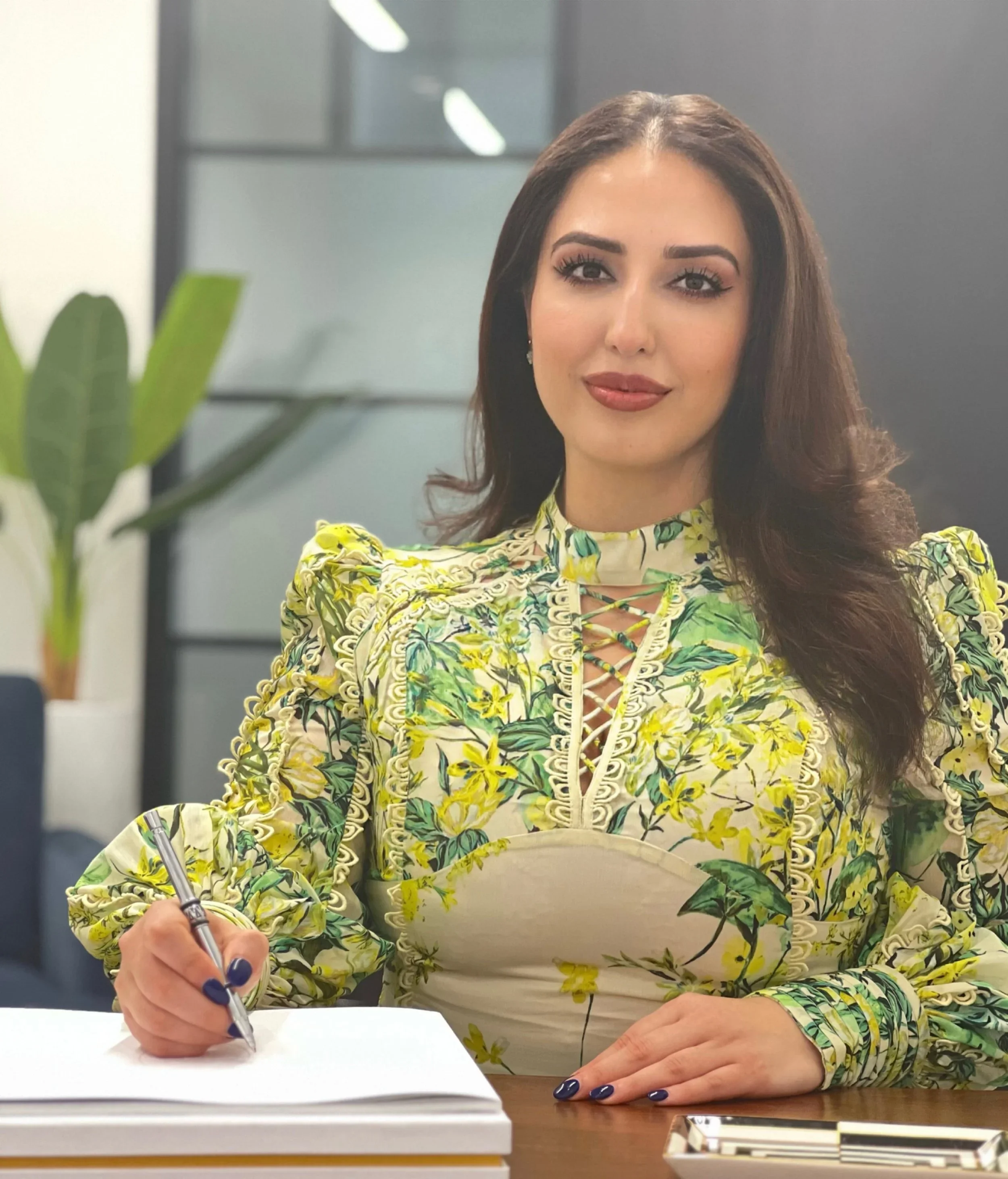 A woman with long dark hair, wearing a yellow floral dress, sits at a table with a pen and notebook, smiling at the camera in a modern office setting.