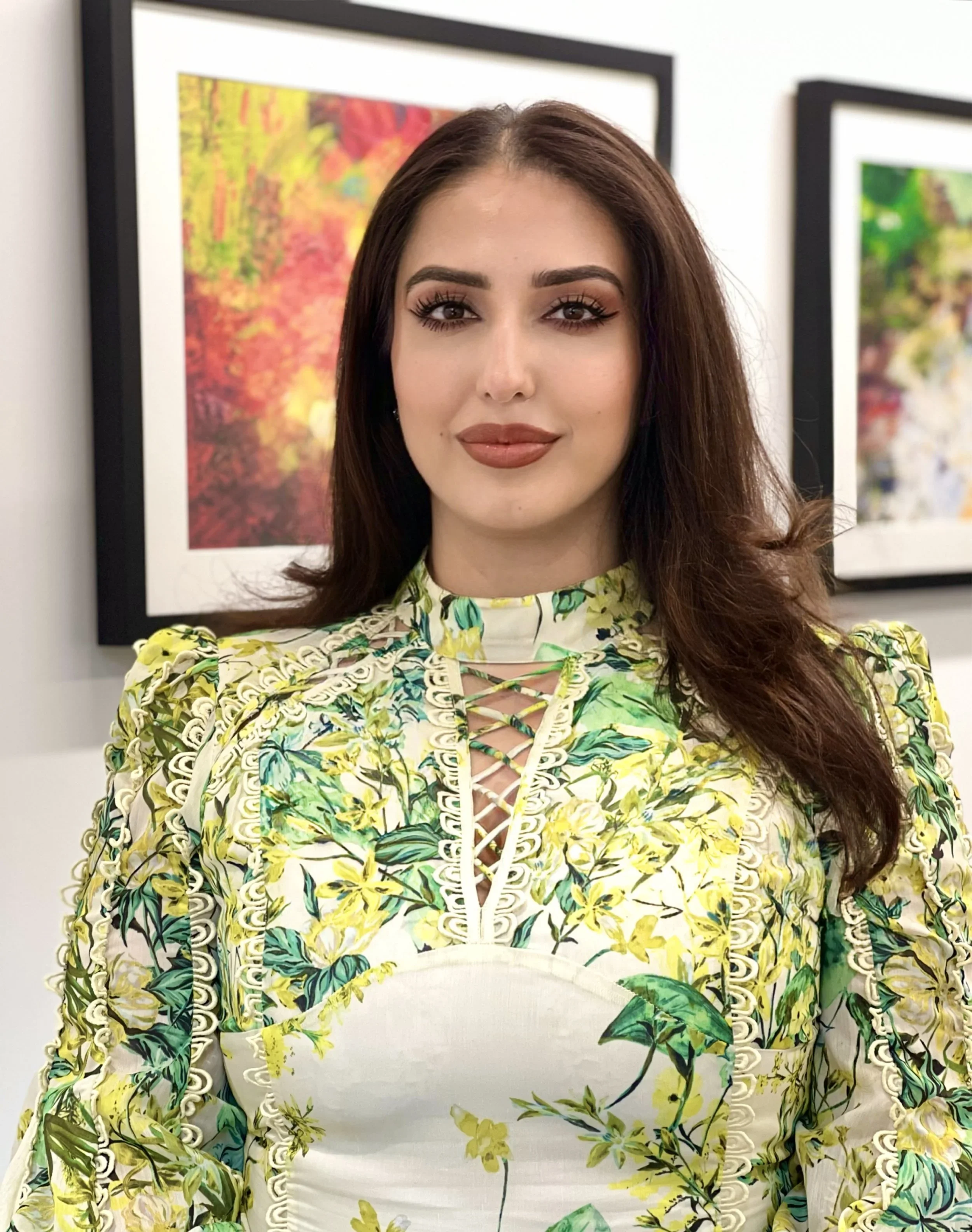 A woman with long brown hair and makeup, wearing a floral patterned dress with green and yellow hues, stands in front of colorful framed art on a gallery wall.