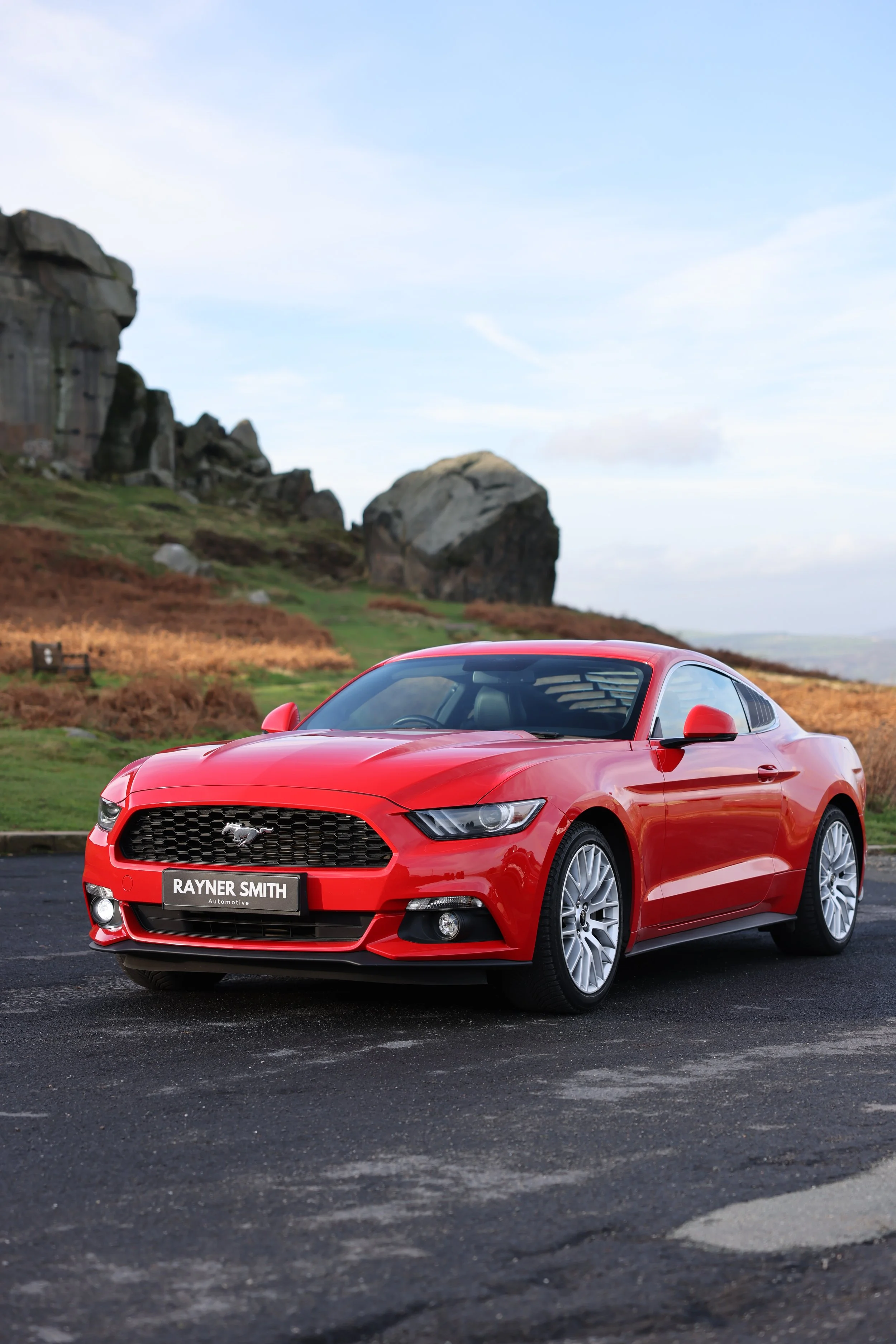 Red Ford Mustang sports car parked on asphalt with rocky hills and cloudy sky in the background.