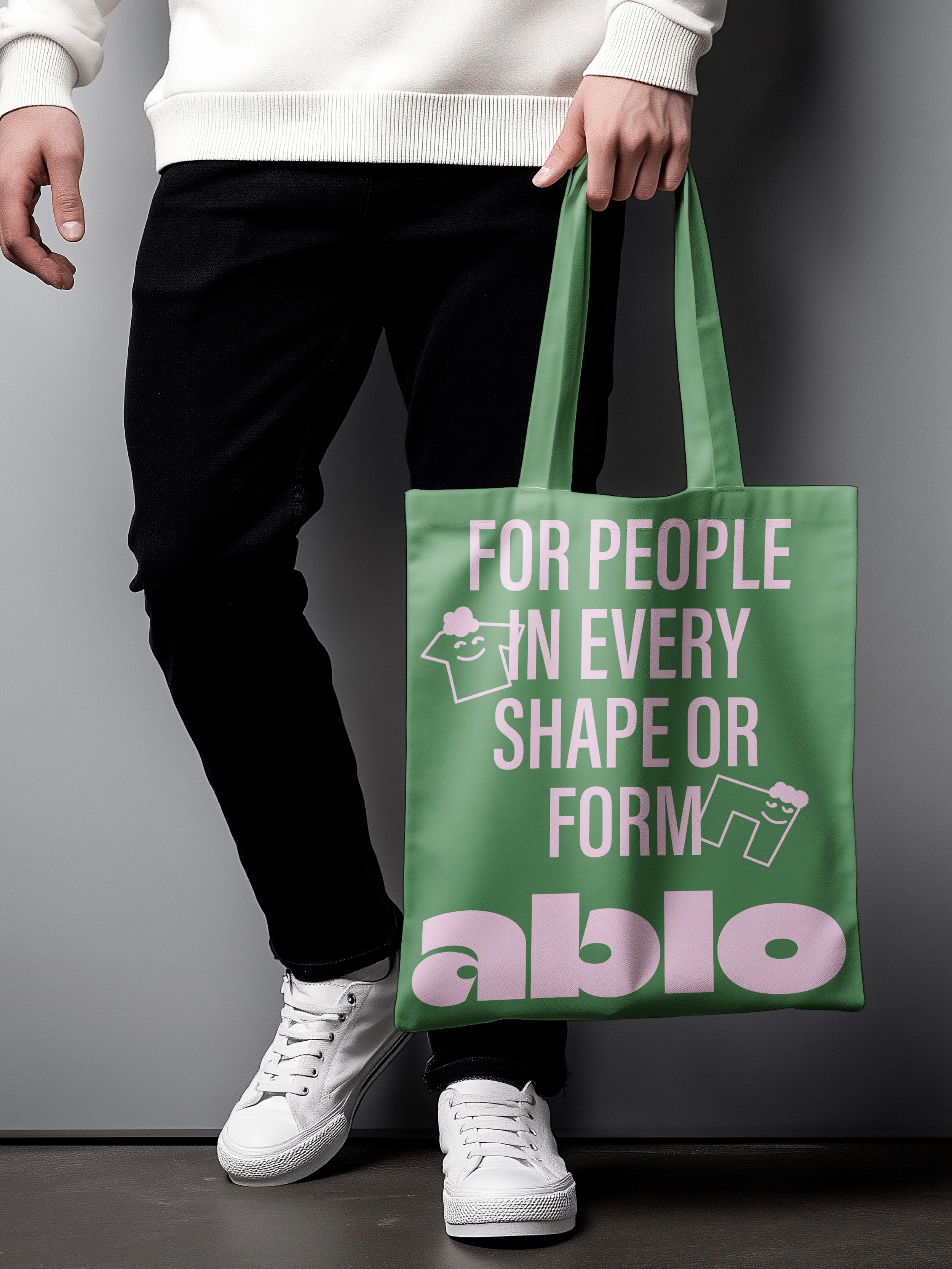 Een persoon houdt een groene totebag met roze tekst vast. De tas heeft de tekst: 'FOR PEOPLE IN EVERY SHAPE OR FORM', en twee leuke figuurtjes. 