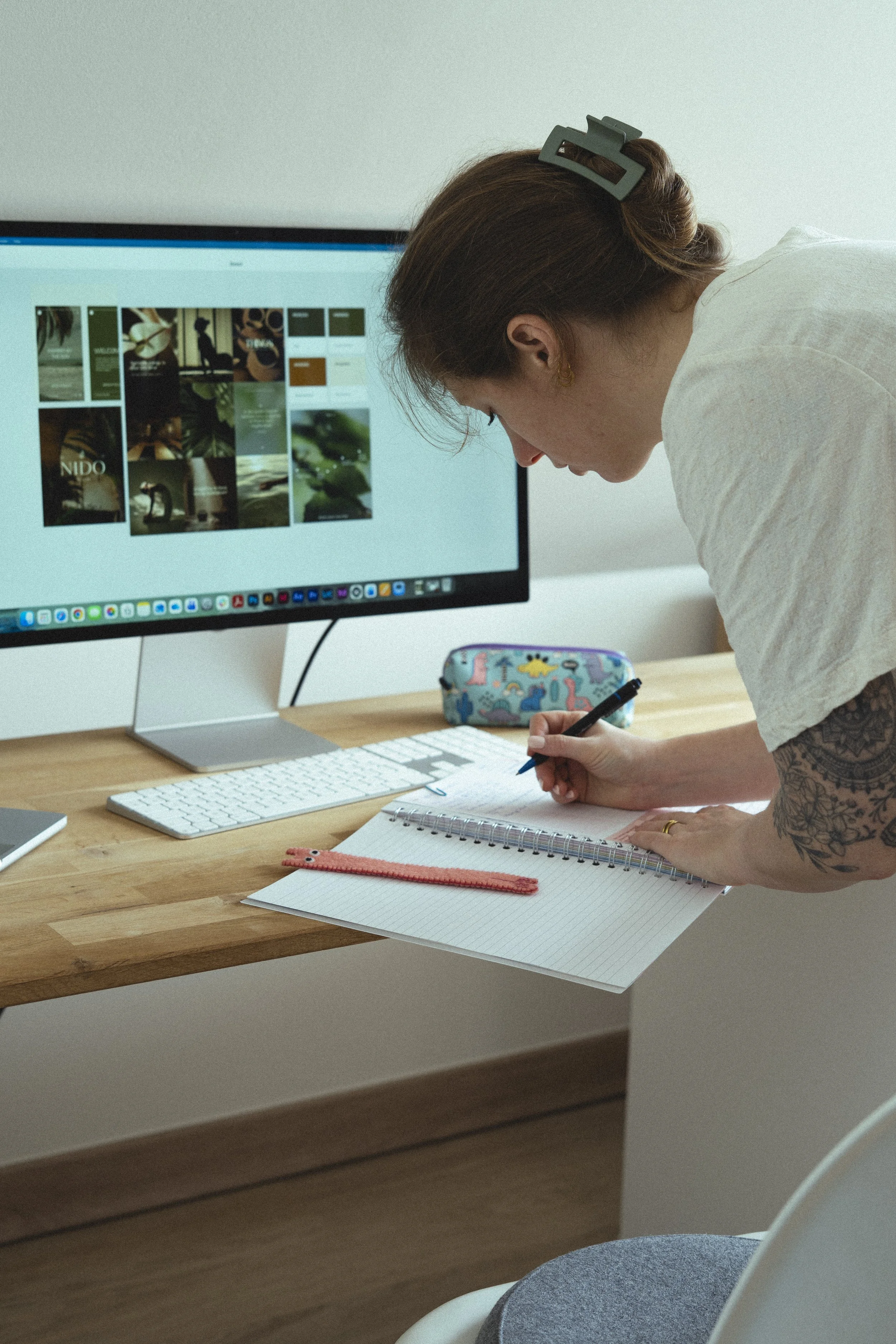 A woman is leaning over her desk, writing in a notebook with a pen. Her workspace includes a desktop computer displaying a collage of images, a white keyboard, a colorful pencil case, and a pink ruler. She has a hair clip, and a tattoo is visible on her arm.