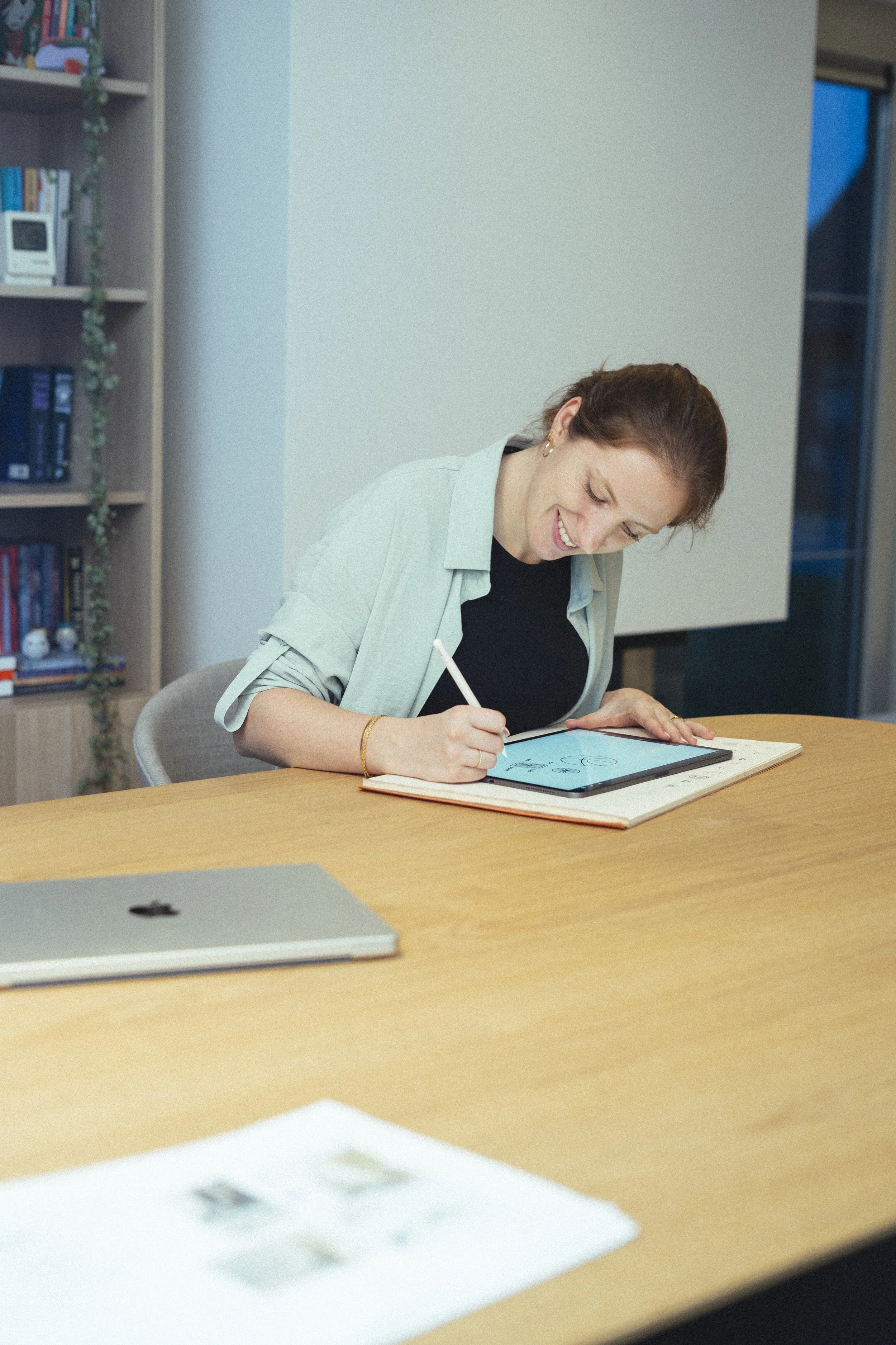 Vrouw zit aan een tafel, werkt aan een digitale tekentablet, glimlacht, met een laptop op de tafel, achtergrond met boekenplank en raam.