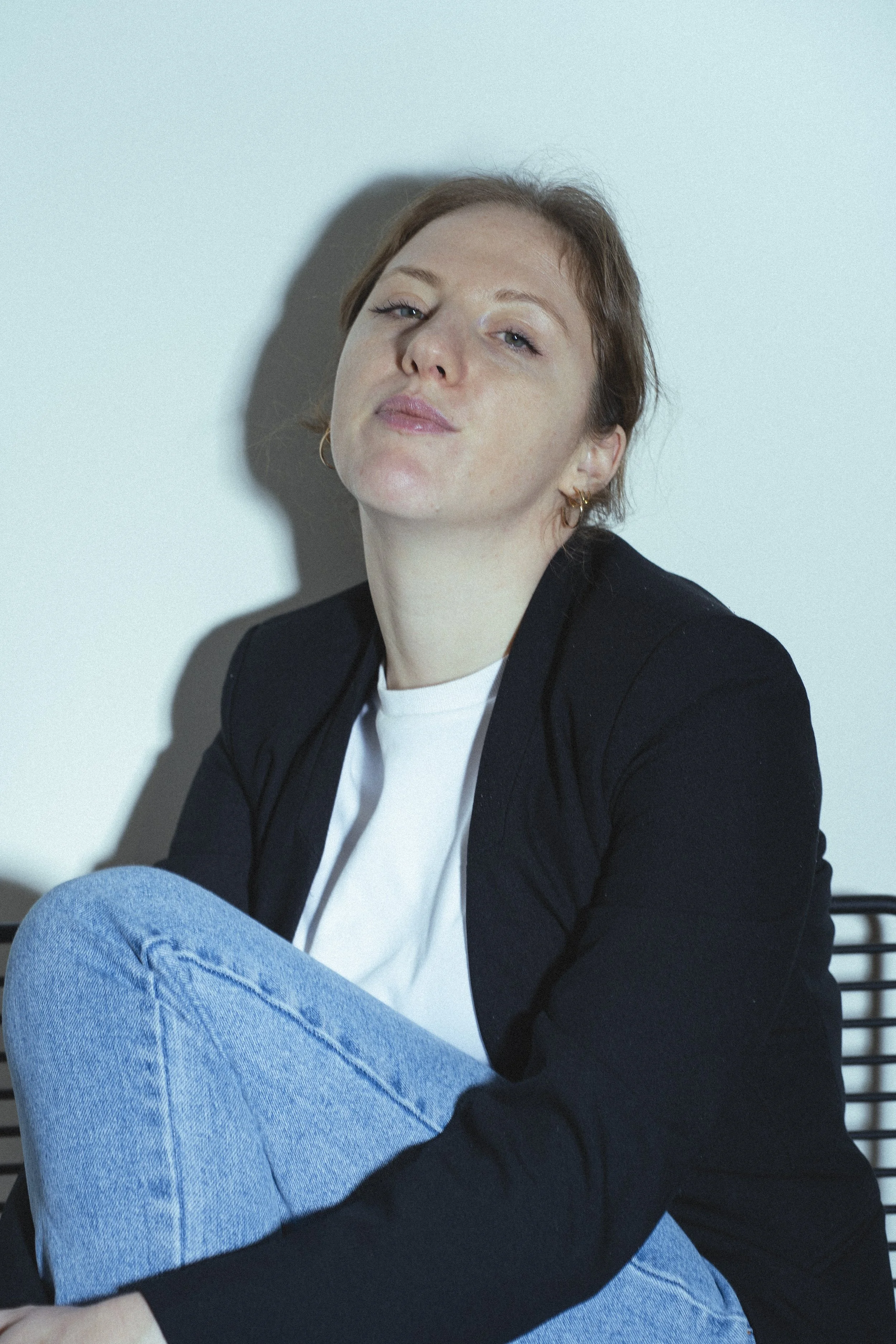 Portret van een jonge vrouw met kort bruin haar, die nonchalant met een lichte glimlach naar de camera kijkt, zittend tegen een lichte muur en gekleed in een zwarte jas en blauwe jeans.