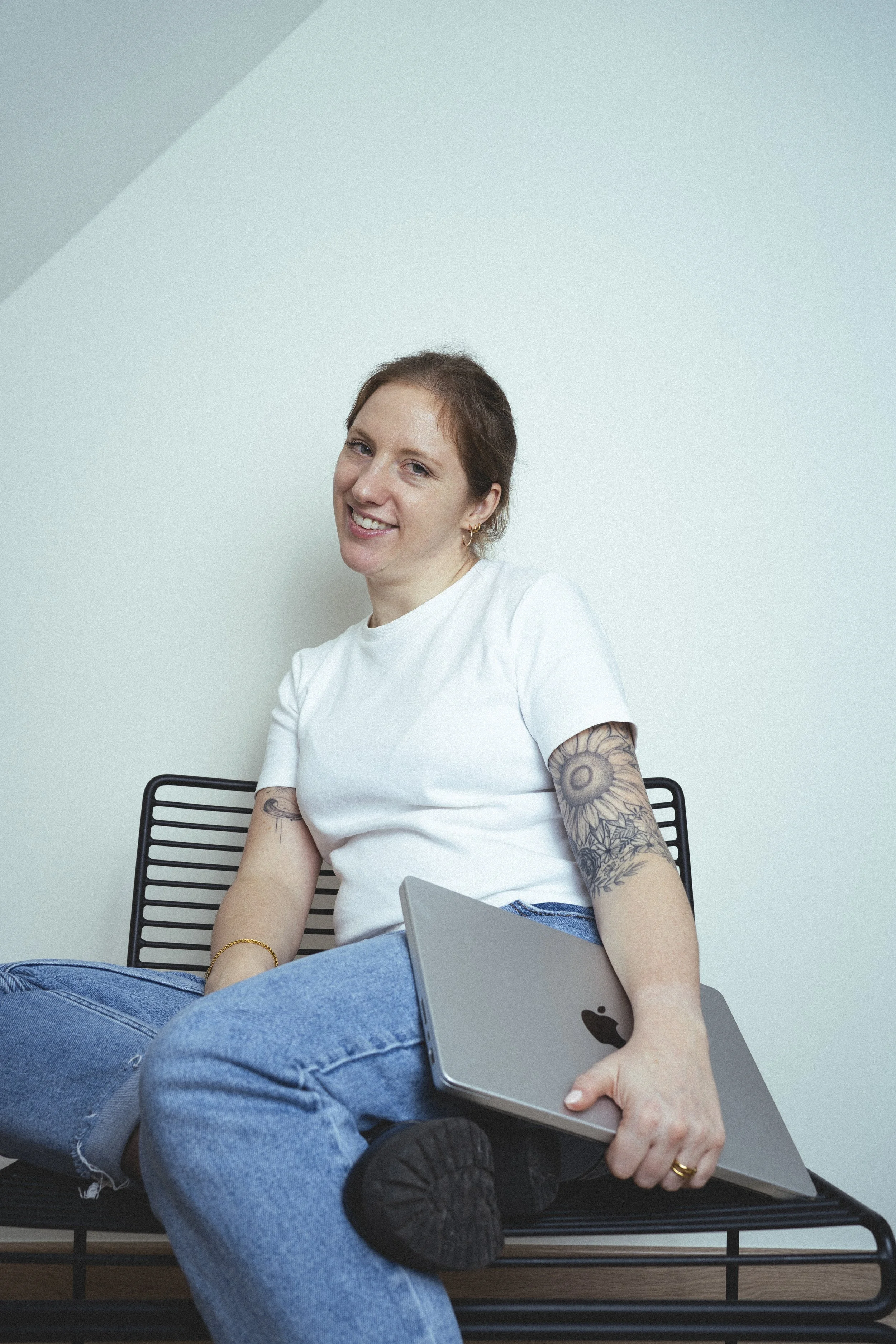 Een vrouw met tattoos op haar armen, gekleed in een witte T-shirt en blauwe jeans, zit op een zwart metalen stoel tegen een lichtblauwe muur. Ze glimlacht en houdt een op laptop rustende hand op haar knie, in haar andere hand een MacBook.