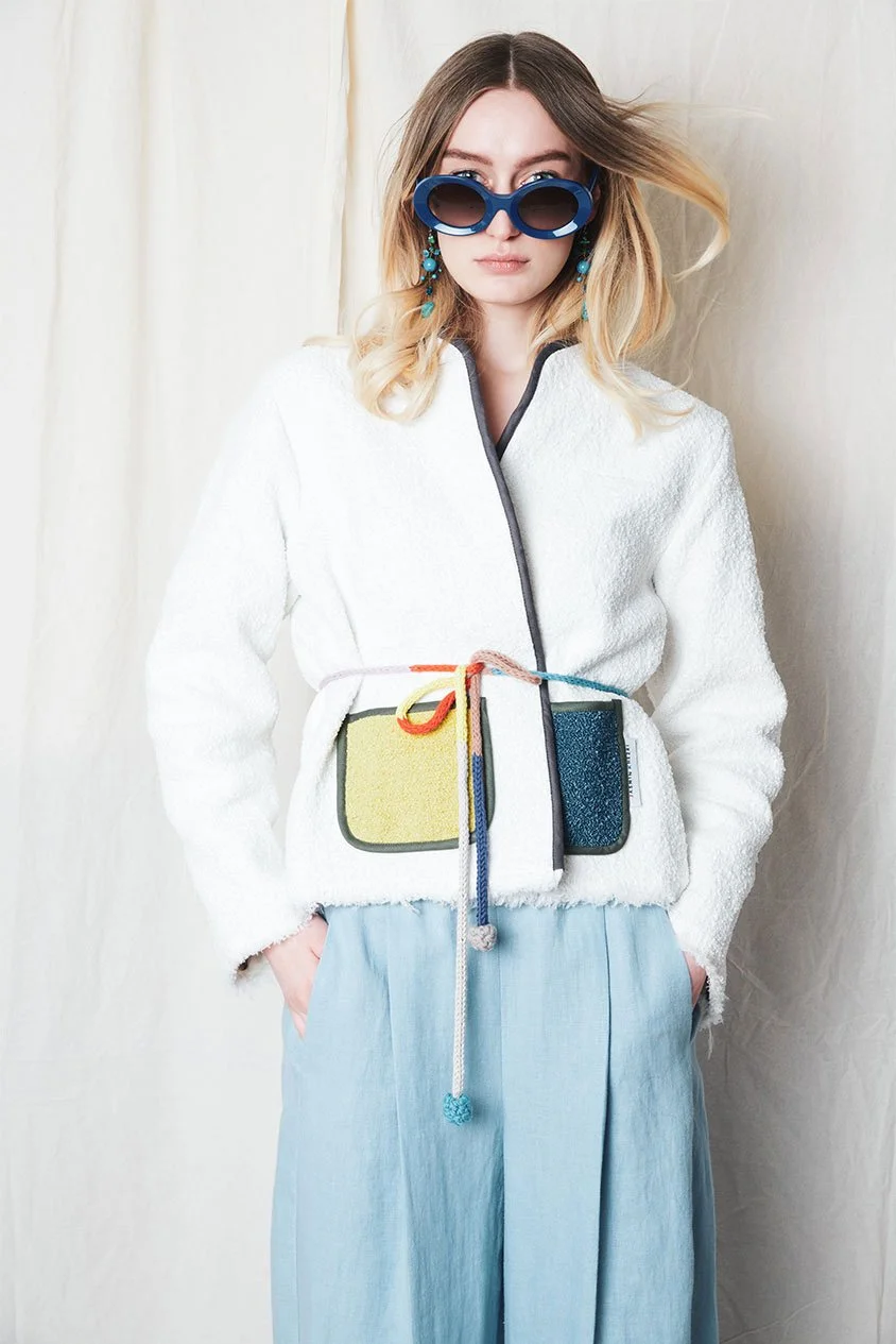 Junge Frau trägt große blaue Sonnenbrille, weiße Jacke, helle Hosen und eine kleine Tasche um den Hals, mit bunten Schnüren