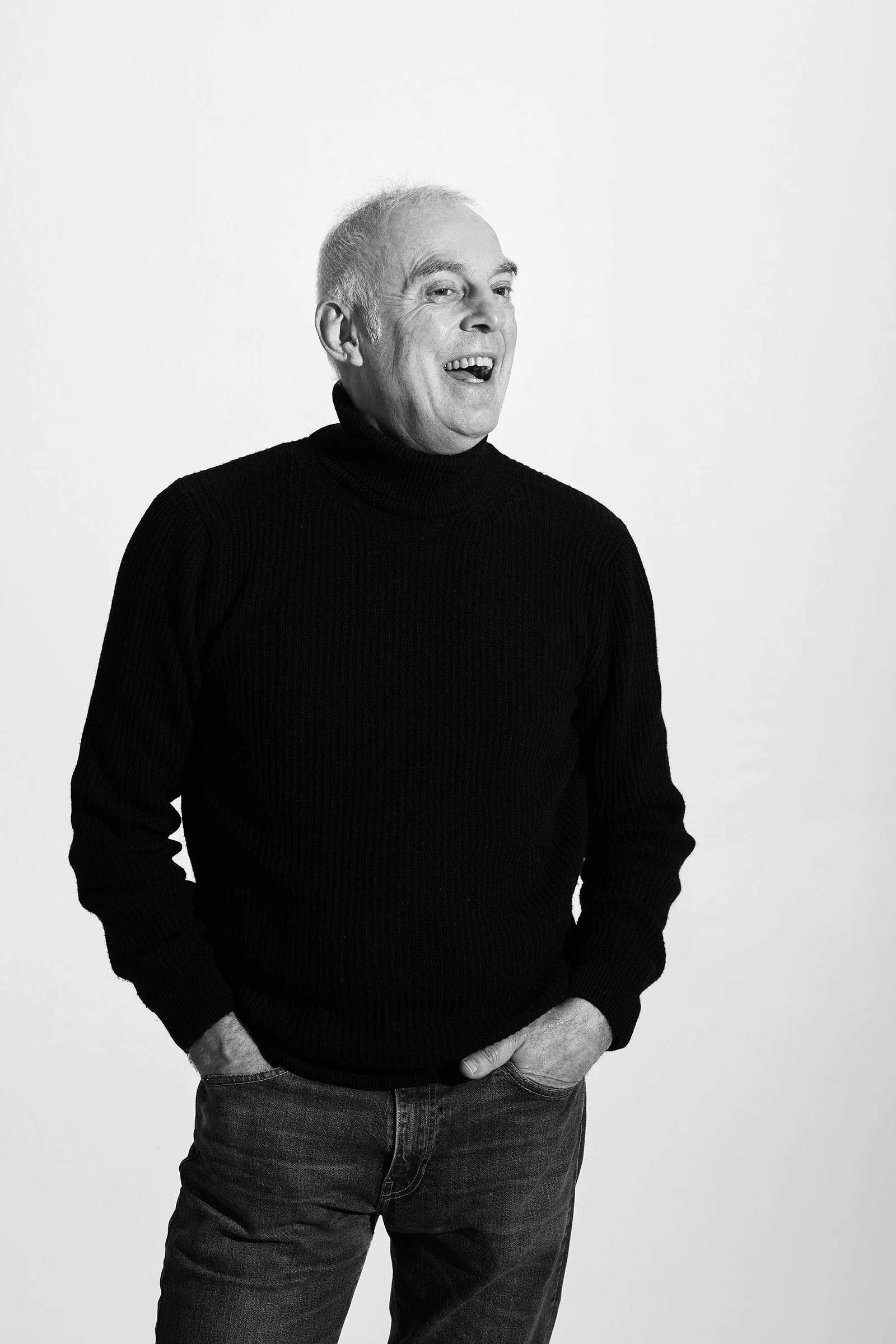 Schwarze und weiße Portraitaufnahme eines älteren Mannes, der lächelnd nach rechts blickt, trägt einen schwarzen Rollkragenpullover und Jeans, vor hellem Hintergrund.