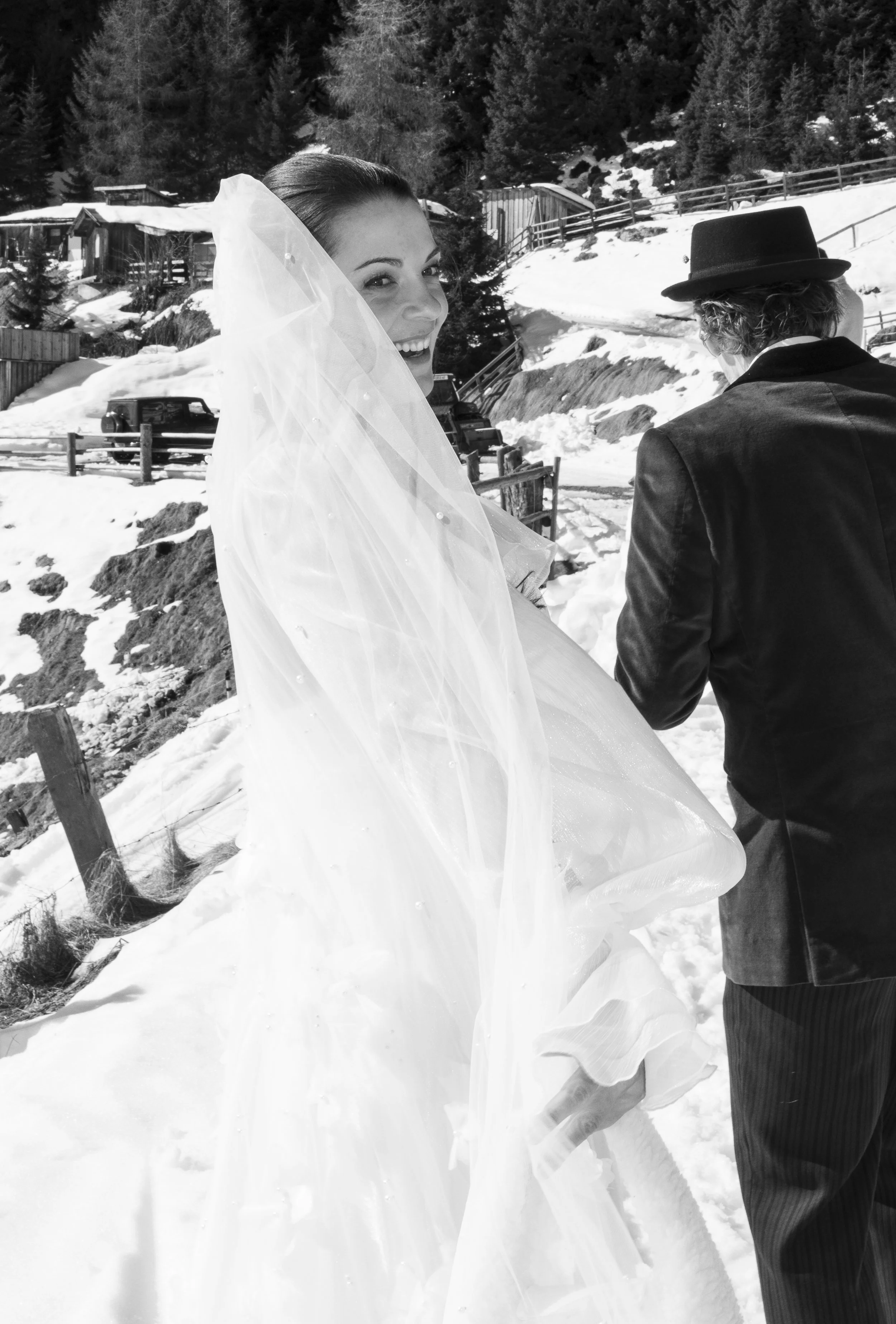Braut in einem weißen Kleid und Schleier blickt lachend in die Kamera, neben einem Mann in dunklem Anzug und Hut in einer verschneiten Berglandschaft.