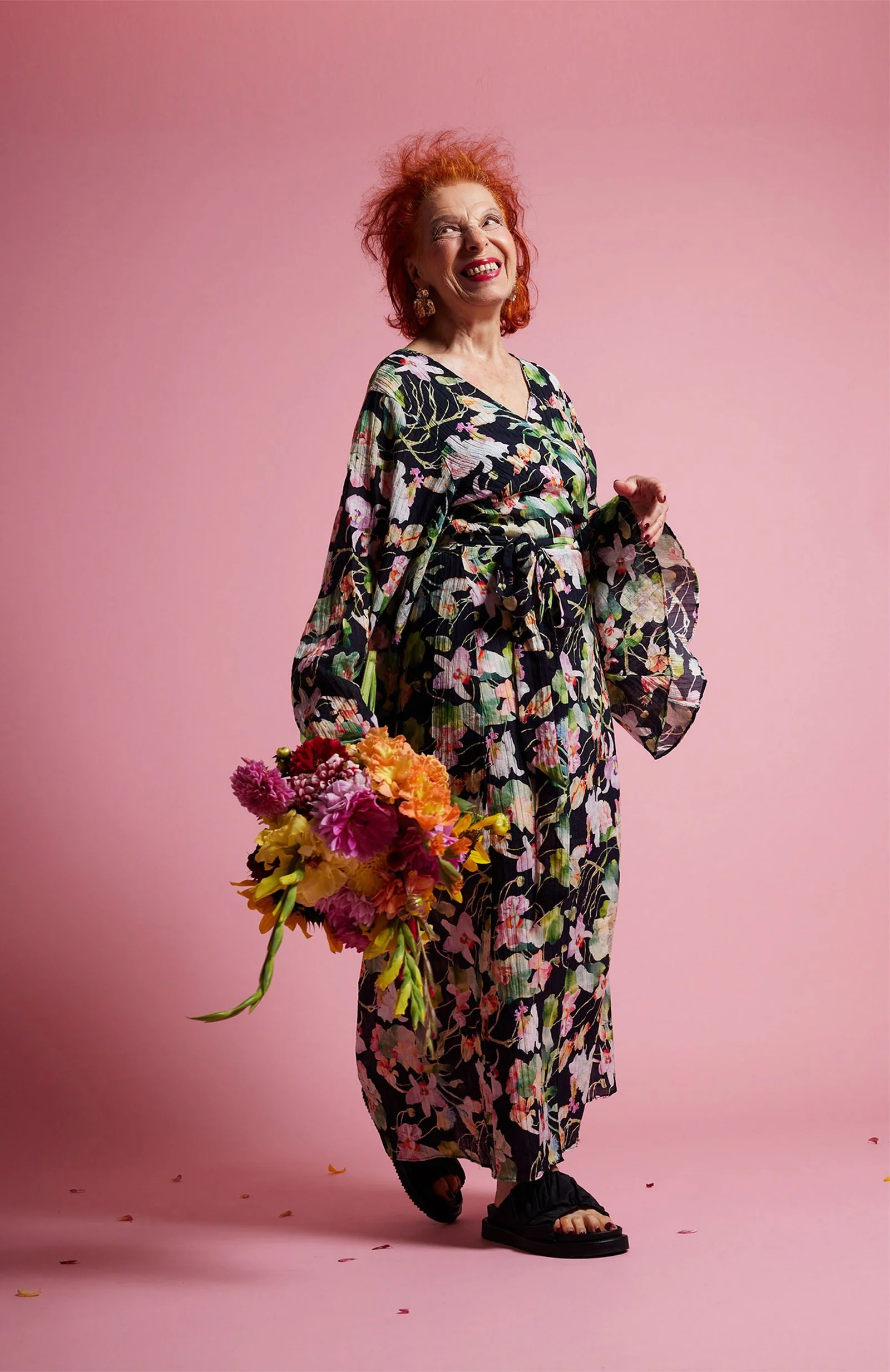 Ältere Frau in einem bunten, floralen Kimono hält einen großen Blumenstrauß und lacht auf einem rosa Hintergrund.