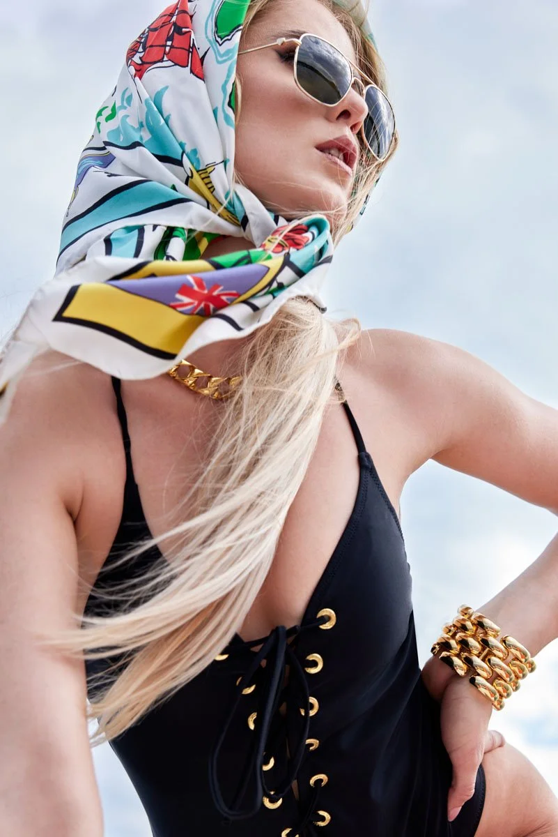 Junge Frau mit Sonnenbrille, bunter Kopftuch, schwarzem Badeanzug, goldener Kette und Armband, im Freien bei Sonnenschein.