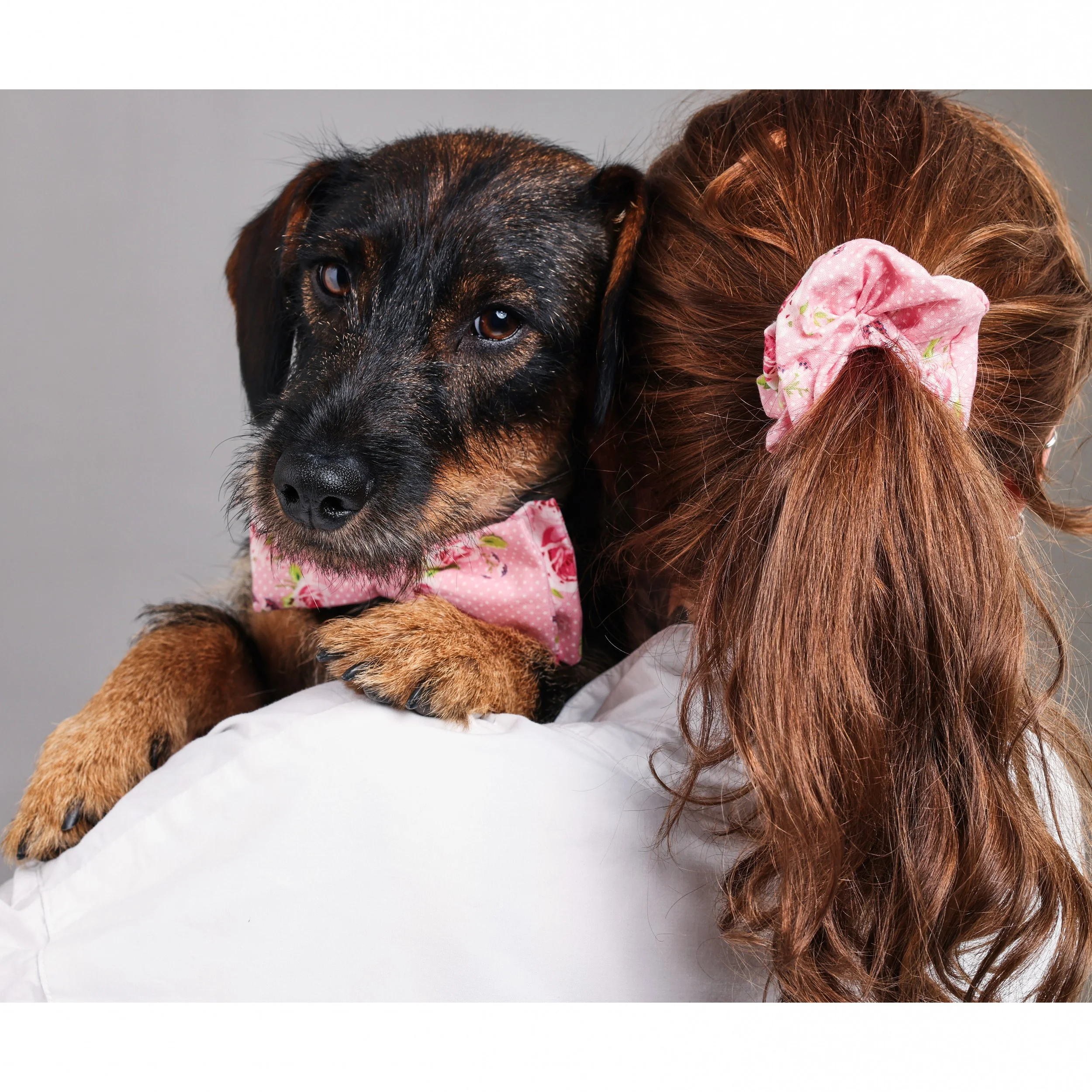 Frau trägt Hund mit braun-schwarzem Fell und rosa Blumenband auf den Schultern.