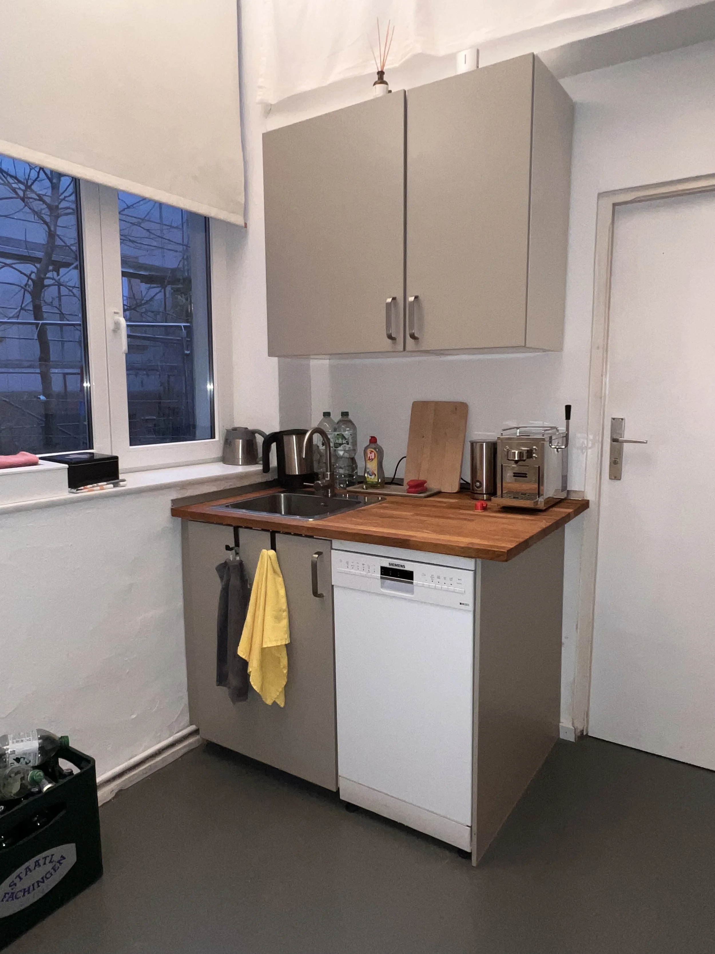 Kleine Küchenecke mit grauen Schränken, Holzarbeitsplatte, Spüle, Wasserkocher, Flaschen und Küchenutensilien, Blick aus dem Fenster auf eine dunkle Außenwelt.