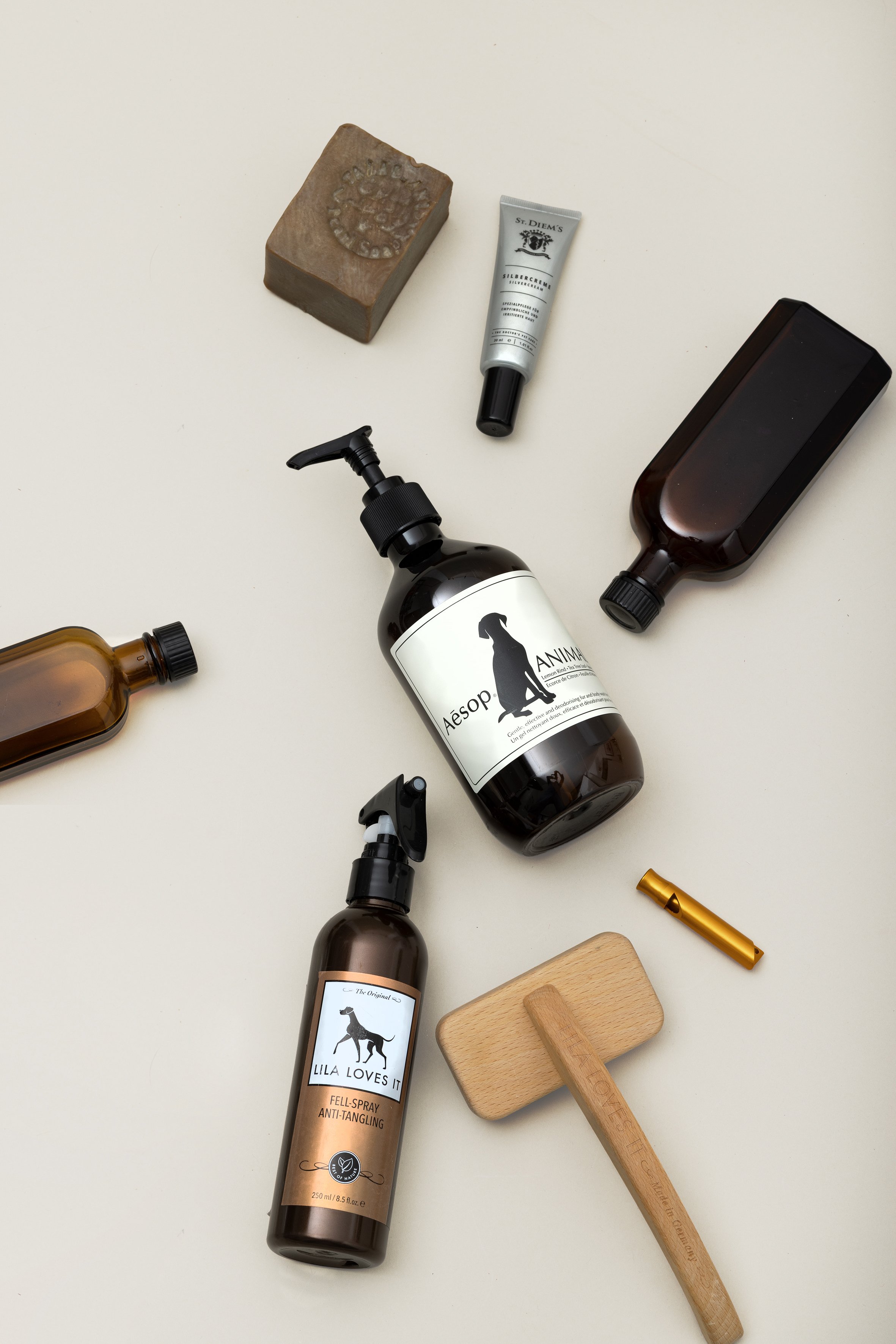 Verschiedene Pflege- und Haushaltsgegenstände liegen auf einem hellen Tisch. Darunter sind Flaschen, ein Stück Seife, ein Holzbrett, ein Holzhammer, ein Spray, ein Spray mit Hundeetikett, ein Pinsel, eine kleine Bürste, ein Tuch und andere Gegenständ