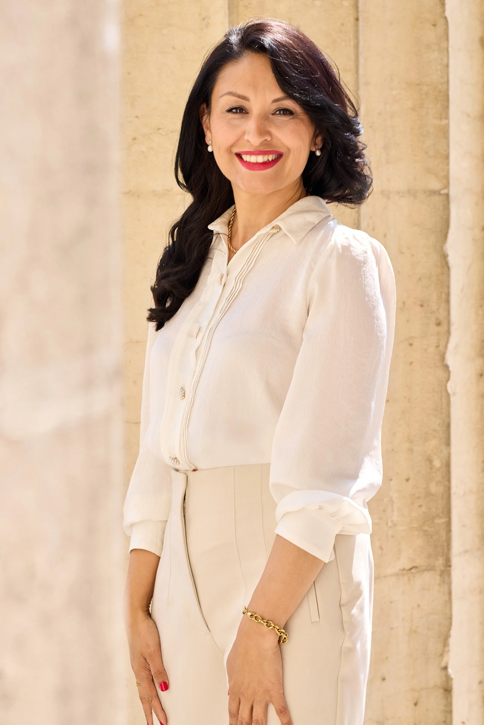 Eine Frau mit dunklen, gewellten Haaren, rot Lippenstift, trägt eine cremefarbene Bluse und Hose, steht vor einer Steinfassade im Sonnenlicht.