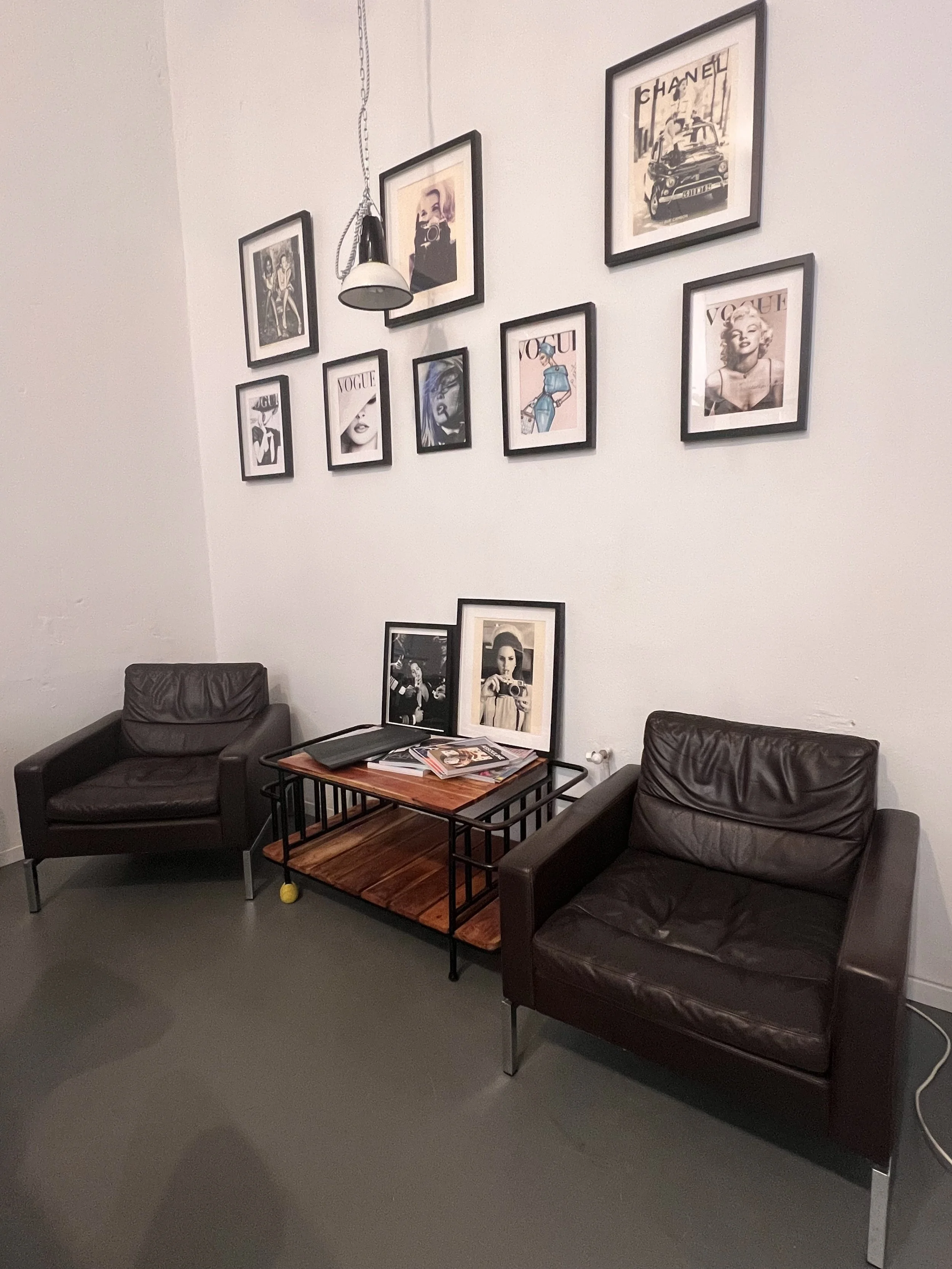 Ein Wartezimmer mit zwei braunen Ledersesseln, einem kleinen Couchtisch mit Zeitschriften und Bildern, und einer Wand mit mehreren Schwarz-Weiß- und Farbfotografien, darunter Vogue-Cover und Modestrecken.
