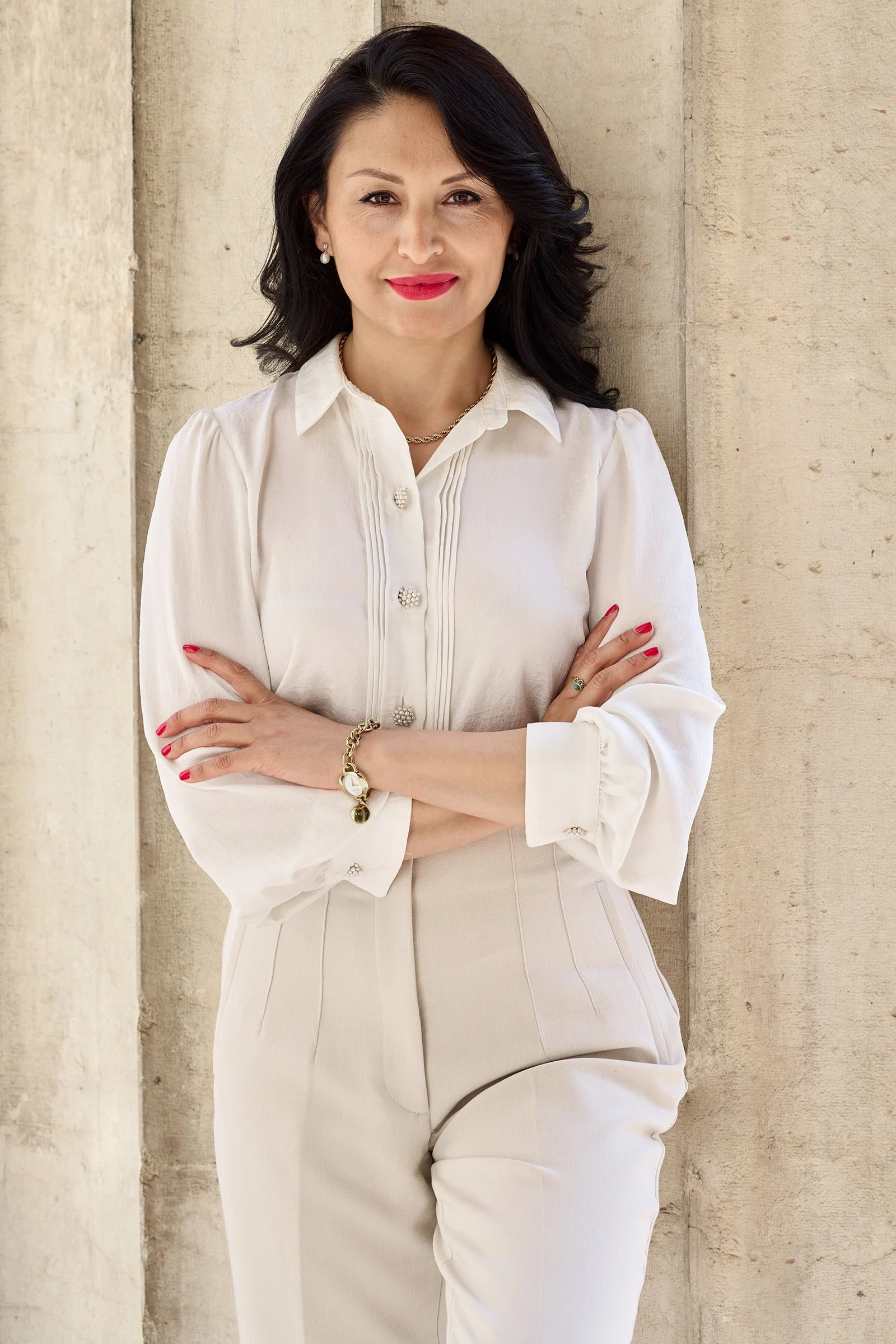 Eine Frau mit dunklen, welligen Haaren, rotem Lippenstift und roten Nägeln, trägt eine weiße Bluse mit Perlmuttknöpfen und cremefarbene Hosen, steht vor einer hellen Steinmauer.