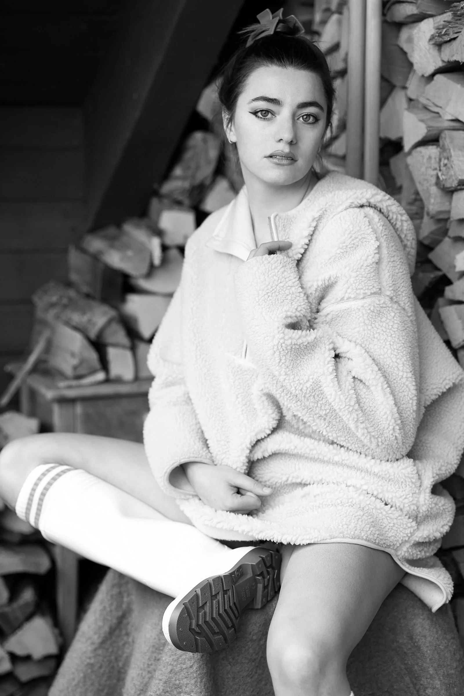 Schwarz-Weiß-Foto eines jungen Mädchens in Pullover, mit Strümpfen und Sneakers, sitzend vor einem Holzstapel.