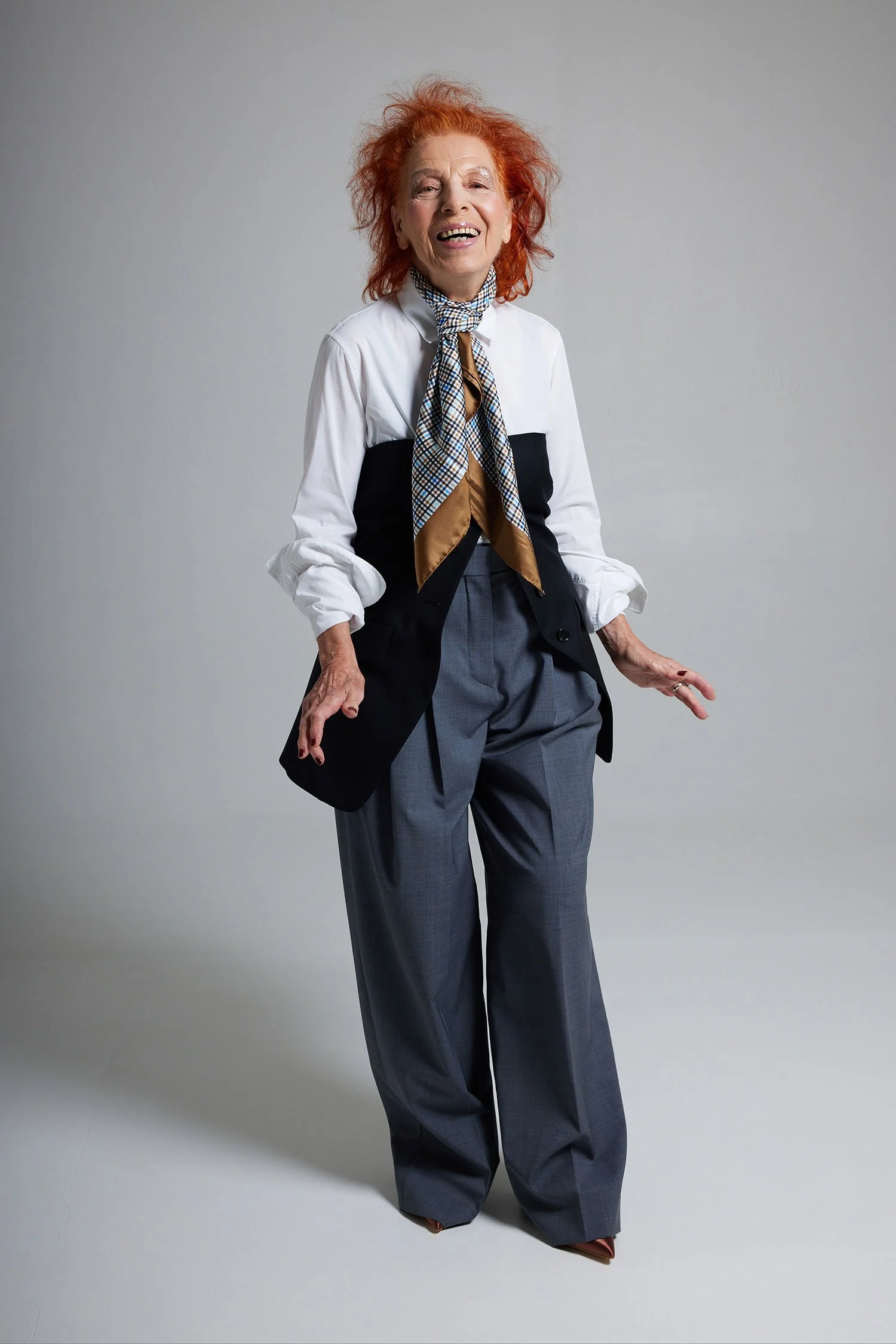 Eine ältere Frau mit roten, lockigen Haaren, lächelnd, trägt eine weiße Bluse, einen schwarzen Blazer mit auffälligen Schultern, eine gemusterte Halskette, breite graue Hose und braune Schuhe.