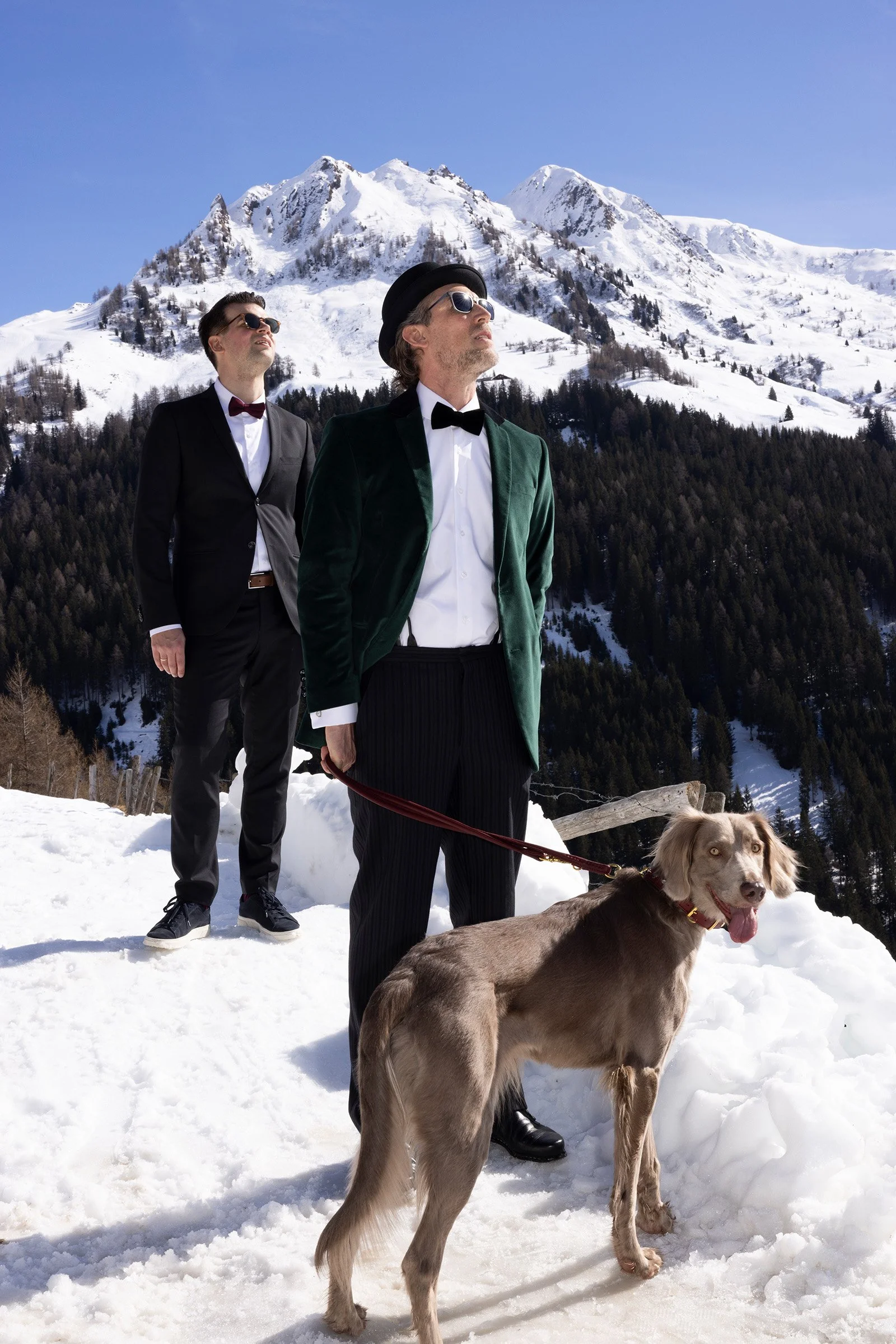 Zwei Männer in formeller Kleidung, einer mit einem Hund an der Leine, auf schneebedecktem Berggipfel mit Schnee bedeckten Bergen im Hintergrund, blauer Himmel.
