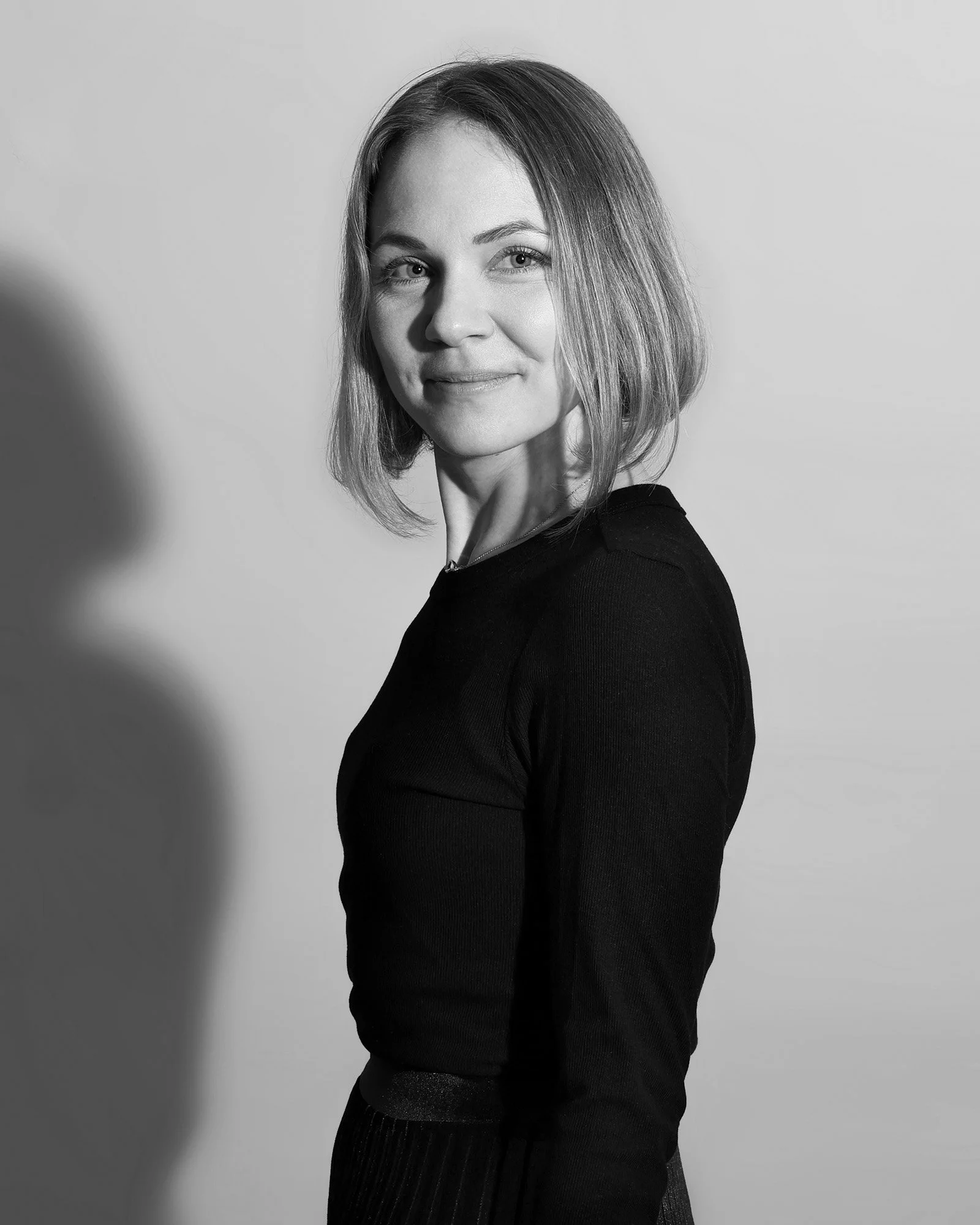 Schwarz-Weiß-Foto einer jungen Frau mit Schulterlänge Haar, die in die Kamera lächelt, vor hellem Hintergrund.
