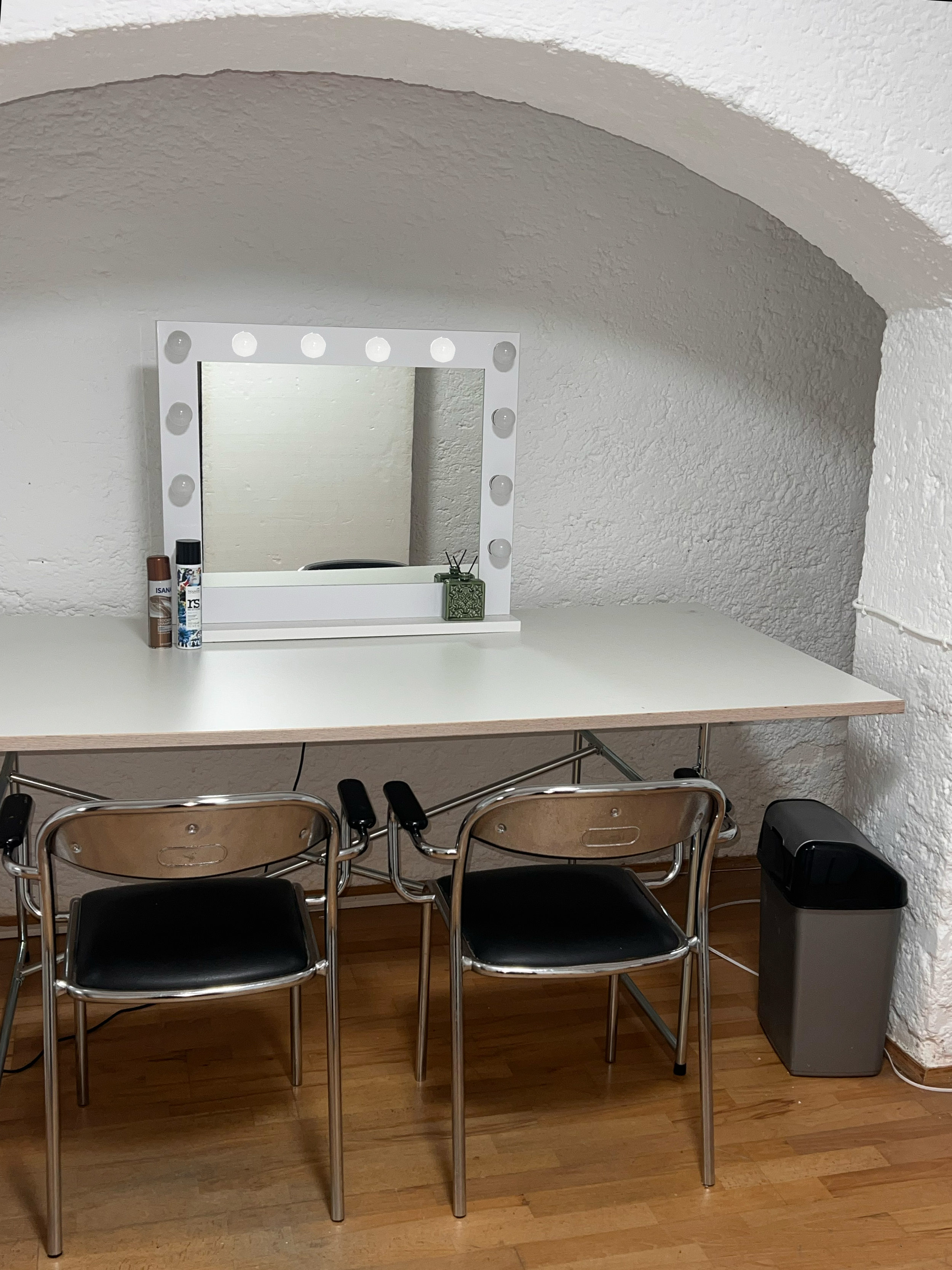 Ein Weißer Schminktisch mit beleuchteter Spiegel, zwei schwarze Stühle, einige Kosmetik- und Pflegeprodukte, eine grüne Dekokiste, eine Mülleimer, alles befindet sich in einem Raum mit weißen Wänden und Holzboden.