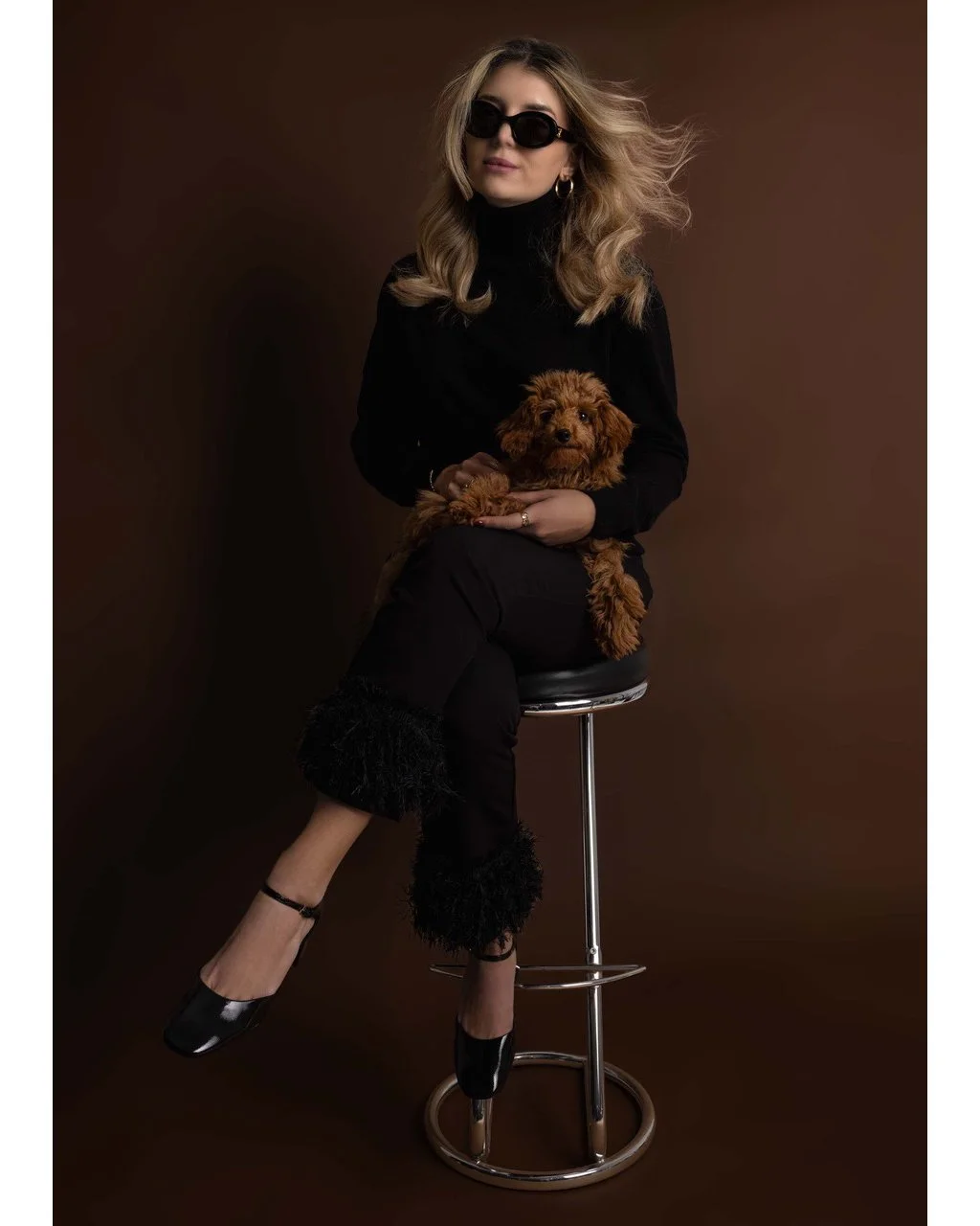 Eine blonde Frau in dunkler Kleidung sitzt auf einem Barhocker und hält einen kleinen braunen Hund auf ihrem Schoß, trägt große Sonnenbrillen und hat lockiges Haar vor einem braunen Hintergrund.