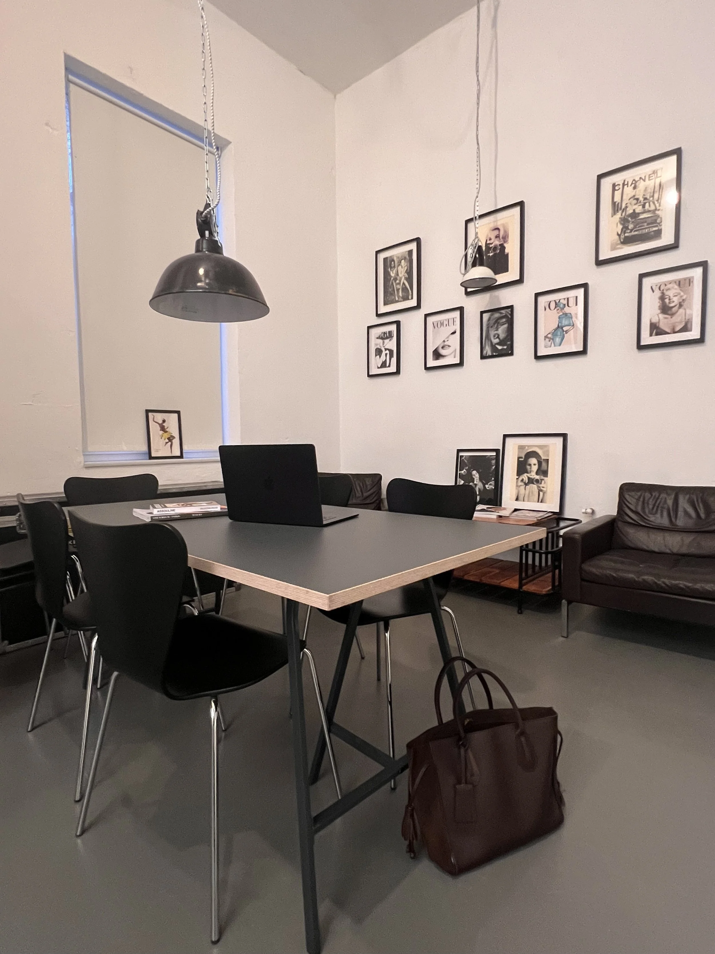 Ein moderner Arbeitsplatz mit einem schwarzen Tisch, sechs schwarzen Stühlen, einem offenen Laptop, einem braunen Ledersessel, einer Tasche auf dem Boden, einer Wand mit mehreren Schwarz-Weiß-Fotomessungen und einem Fenster.