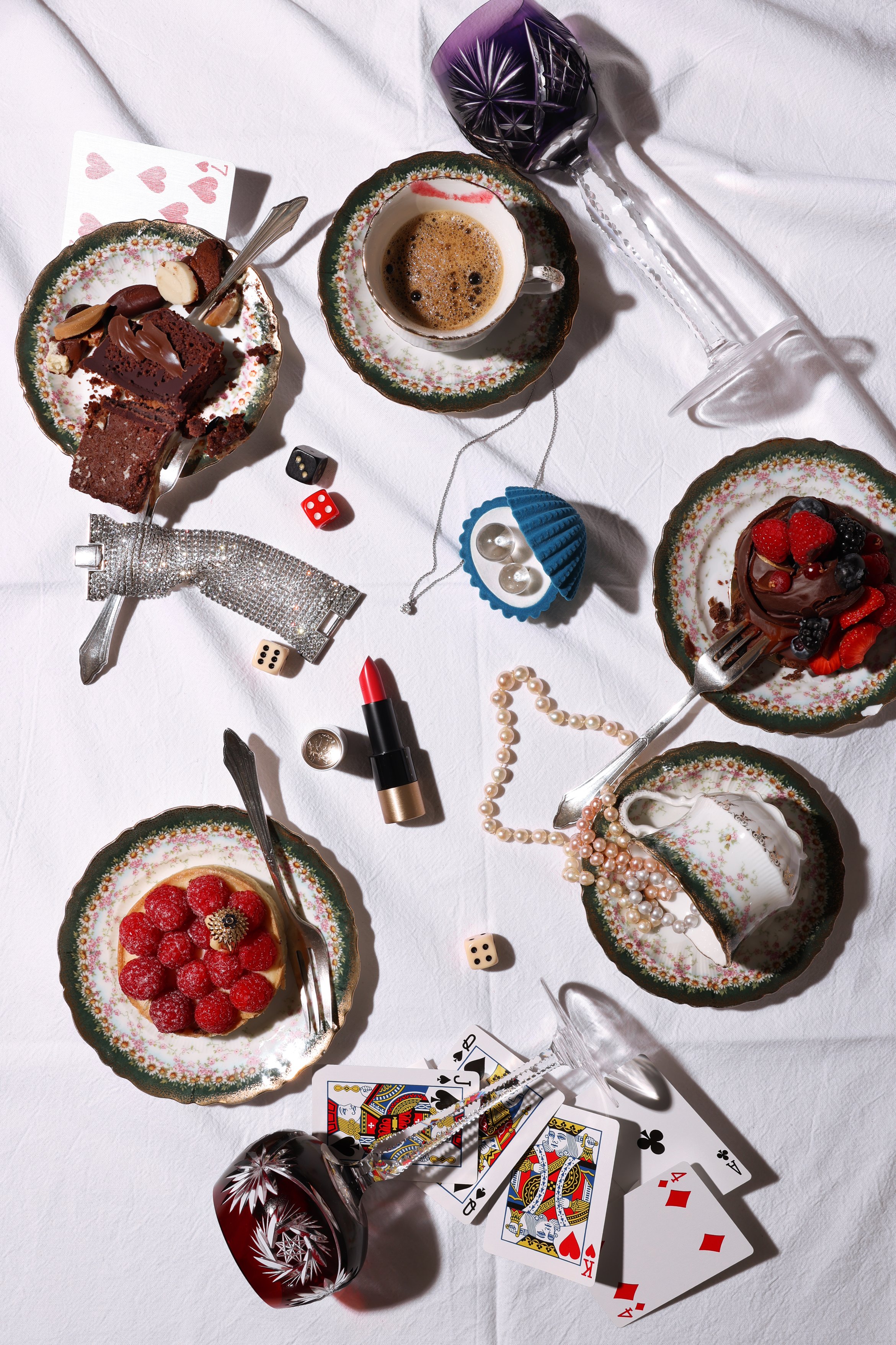 Tisch mit verschiedenen Desserts, Spielkarten, Schmuck, Korkenzieher, Lippenstift, Würfeln und Gläsern, alles auf weißem Tischtuch.