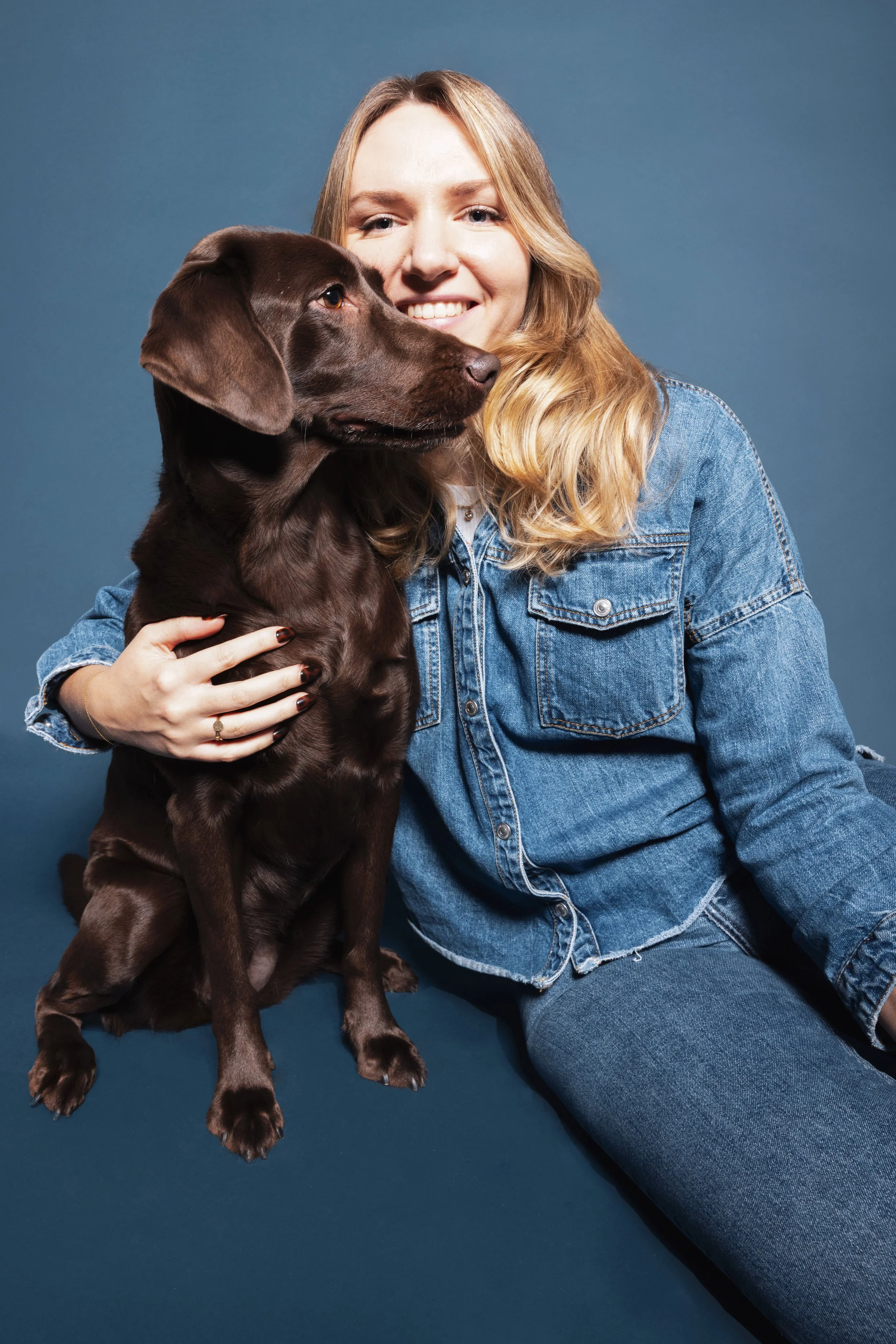 Eine lachende Frau mit langen blonden Haaren, die einen braunen Hund umarmt, vor blauem Hintergrund.