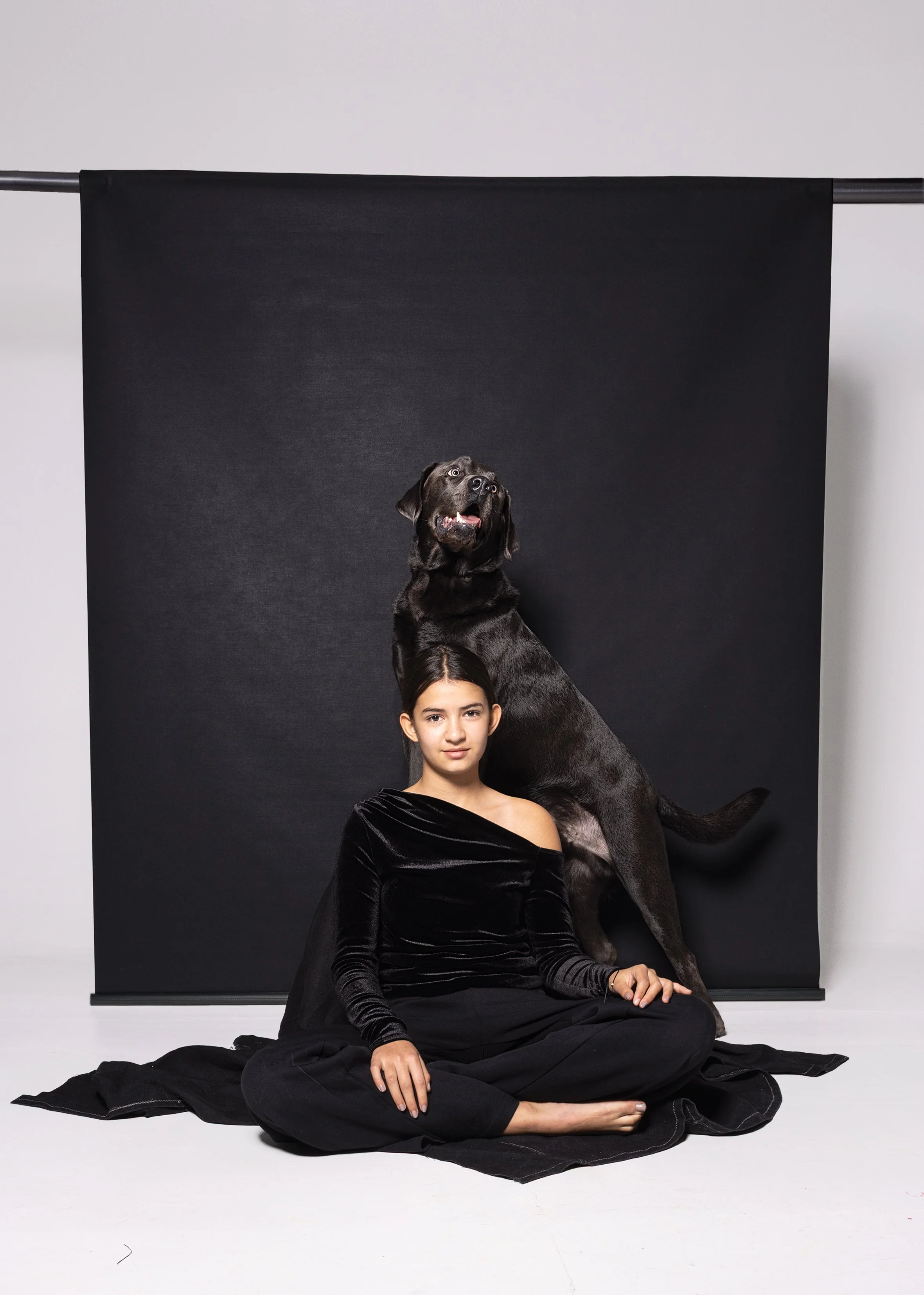 Junge Frau sitzt auf dem Boden in Schwarz vor einem schwarzen Hintergrund, während ein großer schwarzer Hund hinter ihr sitzt.