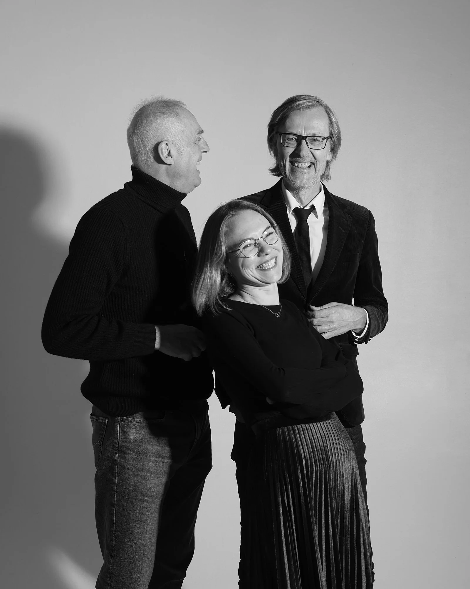 Team Foto in Schwarz-weiß Drei lachende Menschen, zwei Männer und eine Frau, posieren vor einem hellen Hintergrund.