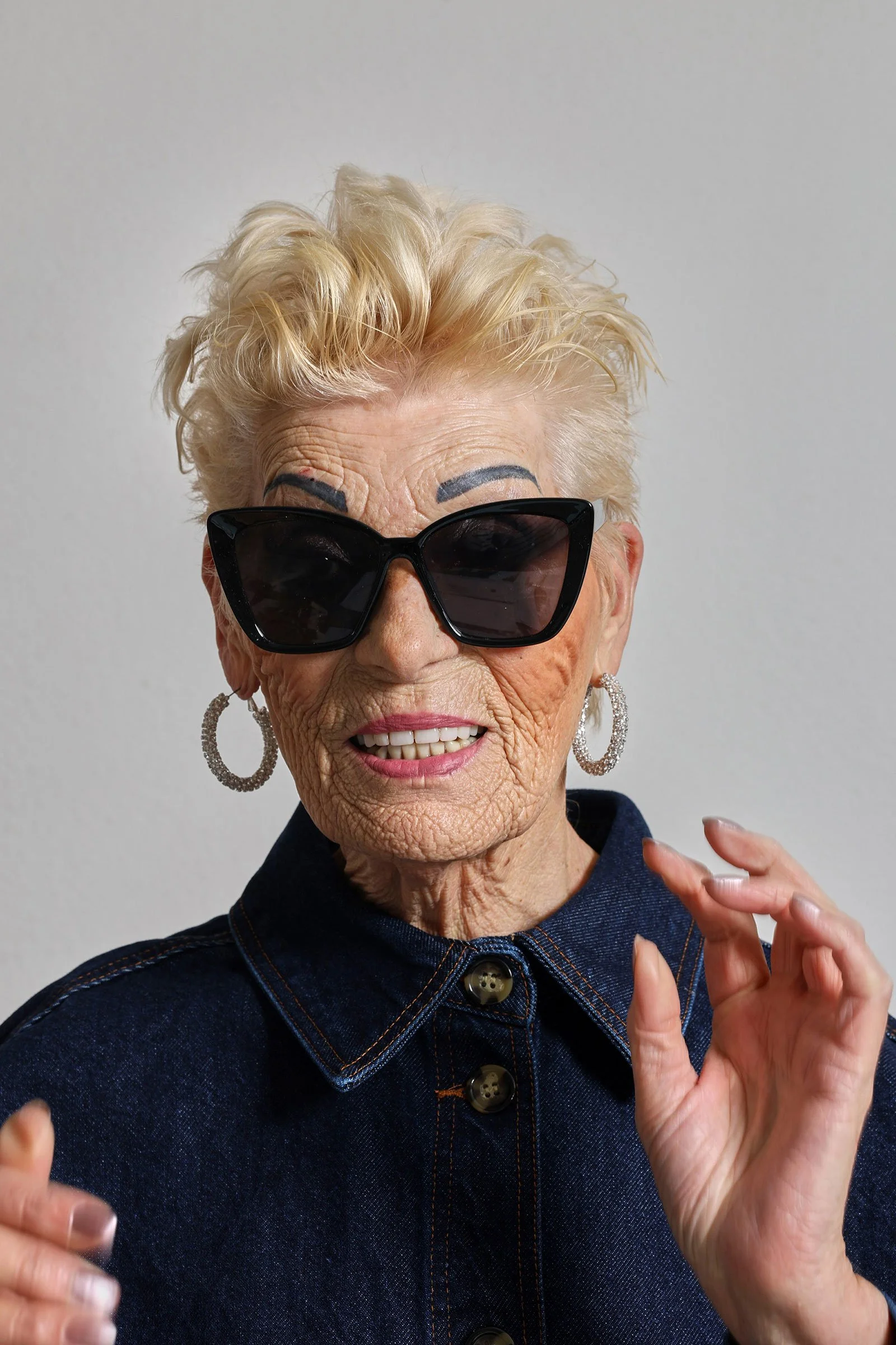 Eine ältere Frau mit blonder, lockiger Kurzhaarfrisur trägt große schwarze Sonnenbrillen, große silberne Ohrringe und eine dunkelblaue Jeansjacke, lächelt freundlich vor einem weißen Hintergrund.