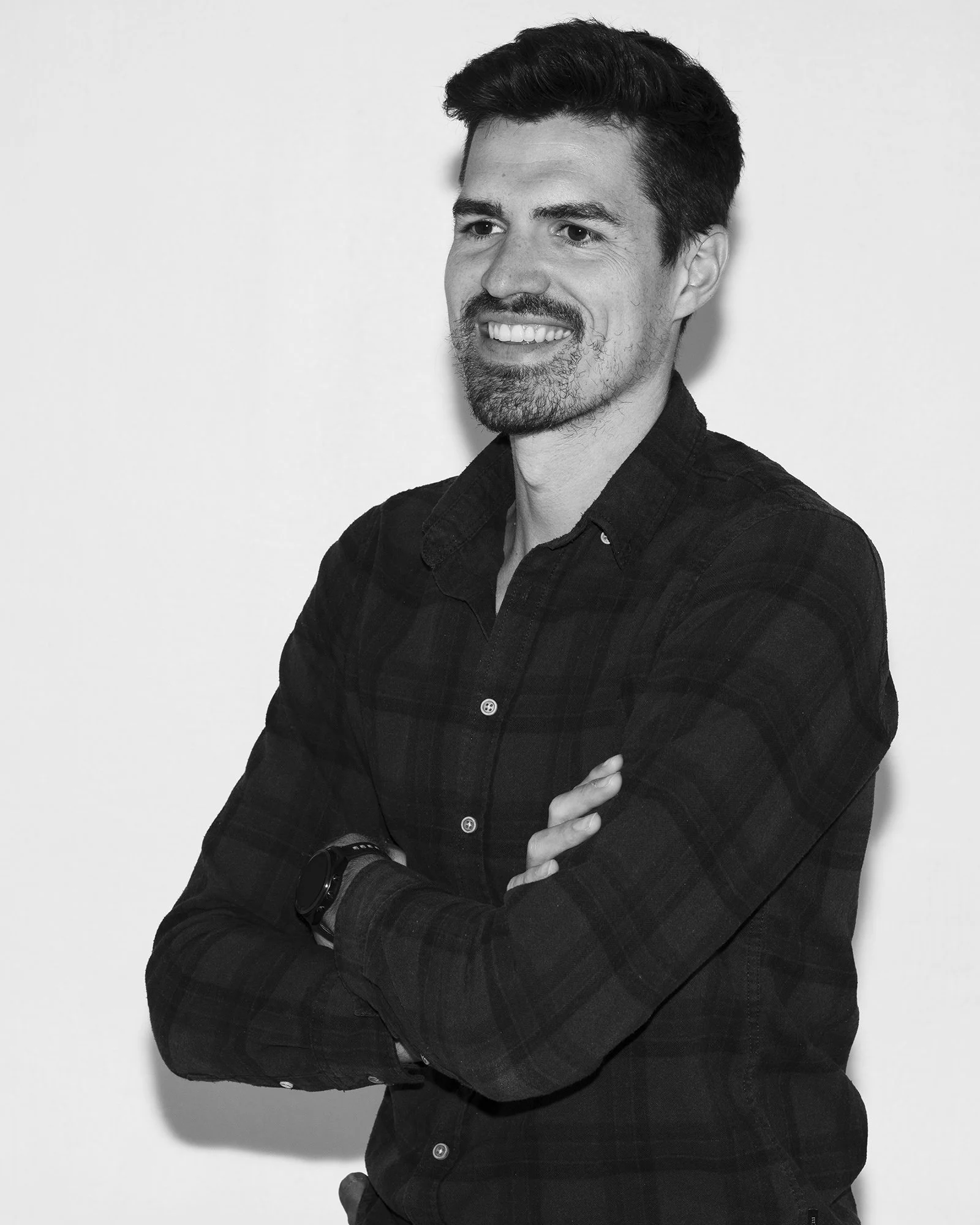 Schwarz-weiß-Portait eines Mannes mit dunklem Haar und Bart, lächelnd, trägt Hemd mit aufgekrempelten Ärmeln, steht vor weißem Hintergrund