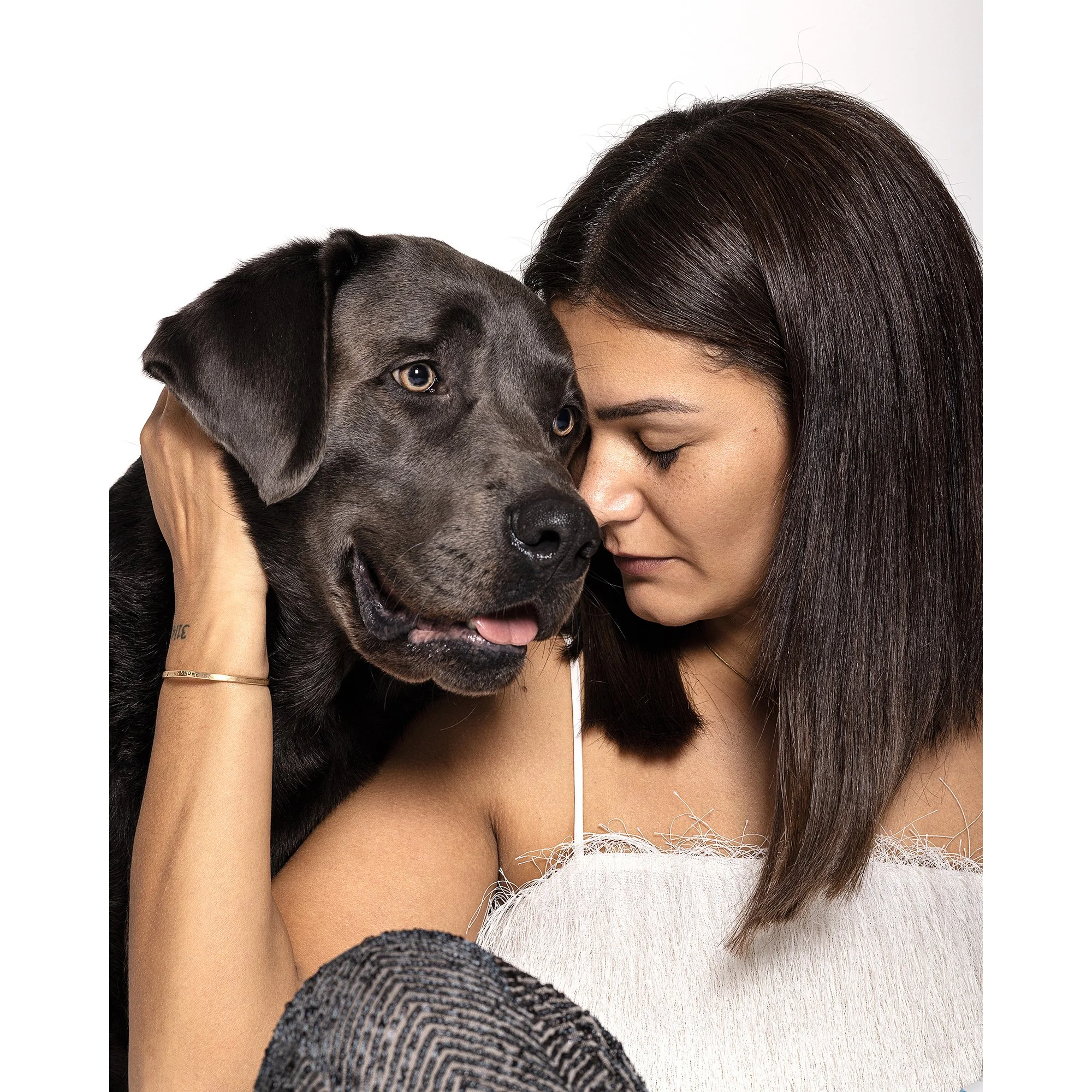 Eine Frau hält einen schwarzen Hund nah an ihr Gesicht, beide haben die Stirn aneinandergelegt, in einem weißen Hintergrund.