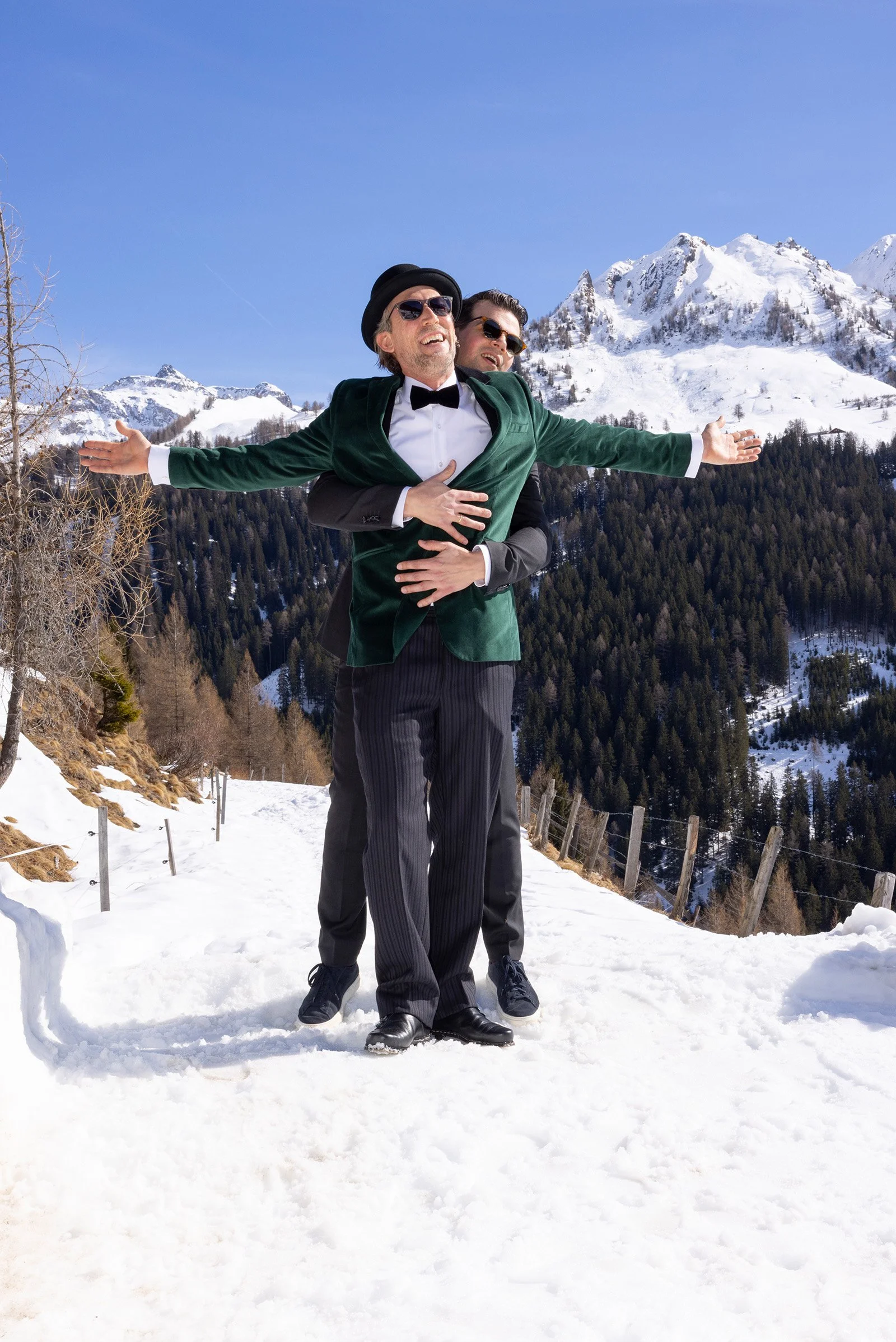 Zwei Männer in formeller Kleidung im Schnee vor verschneiten Bergen, einer umarmt den anderen von hinten, beide lachen.