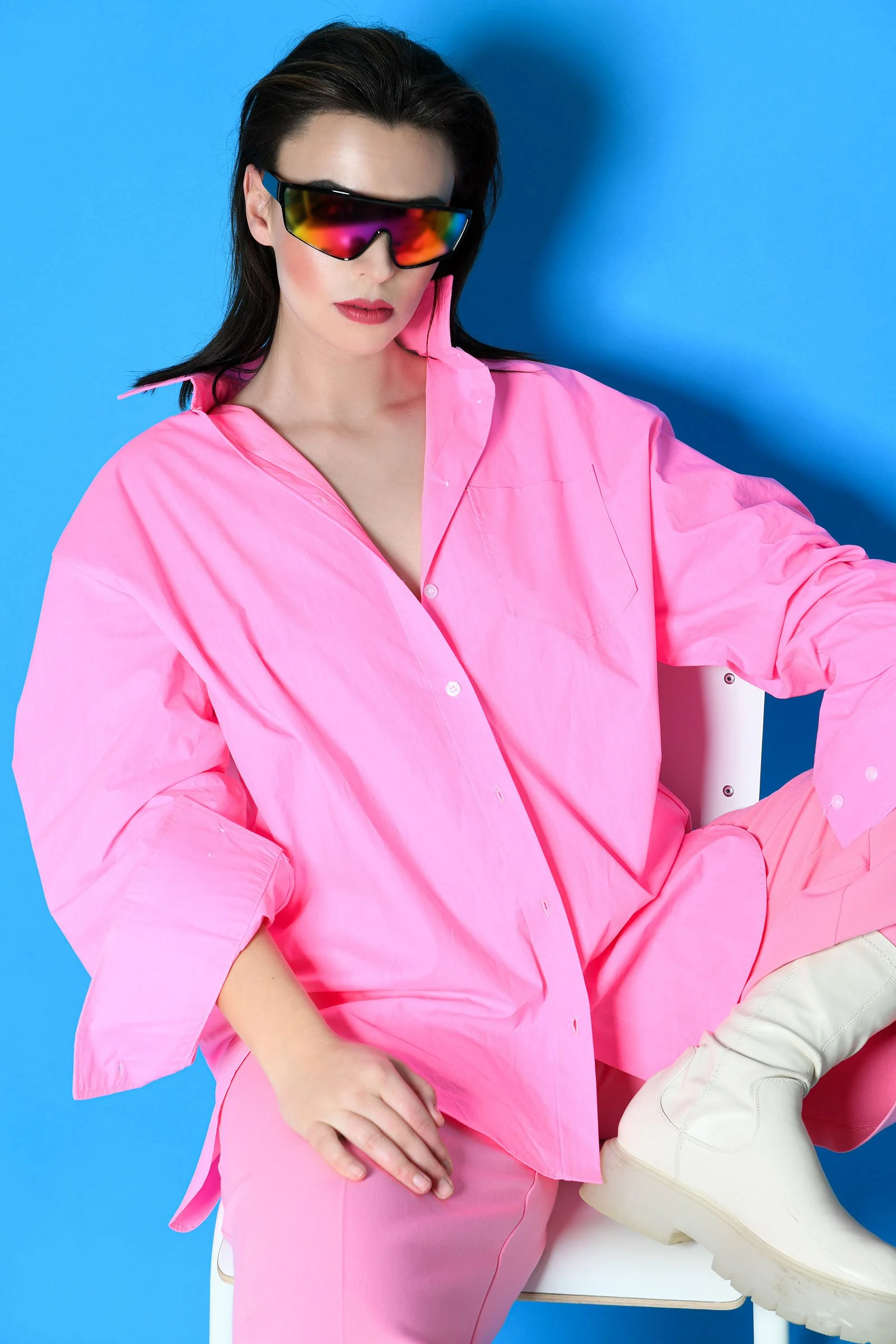 Frau in pinkem Outfit mit schwarzen Sonnenbrillen sitzt auf einem Stuhl vor einem blauen Hintergrund