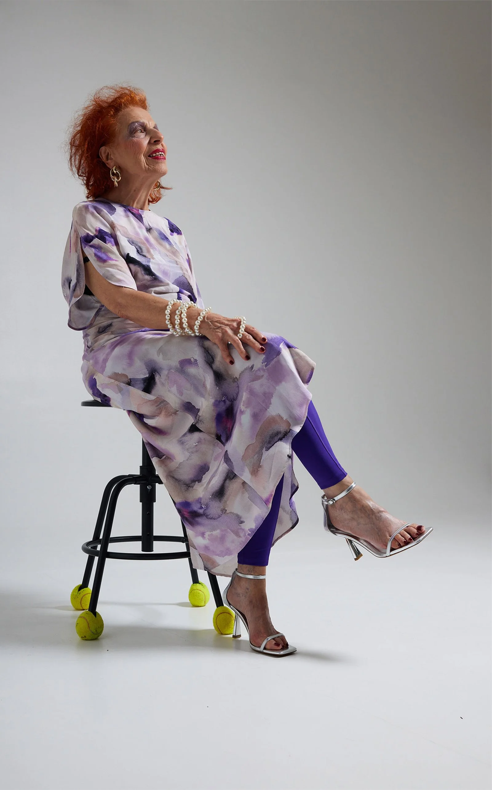Eine ältere Frau mit roten Haaren sitzt auf einem Stuhl, trägt einen bunten, lila-weiß gemusterten Rock, Perlenarmbänder, elegante Schuhe, hat fröhliches Make-up und lächelt, während sie aufrecht sitzt.