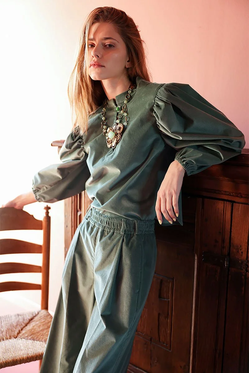 Junge Frau in einer grünen Bluse und hose, mit auffälliger Kette, lehnt an einem Holzschrank in einem Raum mit warmem Licht.