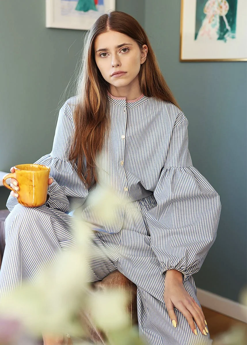 Junge Frau mit langen braunen Haaren sitzt in einem Raum, trägt einen gestreiften Pyjama und hält eine gelbe Tasse in der Hand. Sie sieht ernst oder gelangweilt aus, im Hintergrund sind Bilder an der Wand.