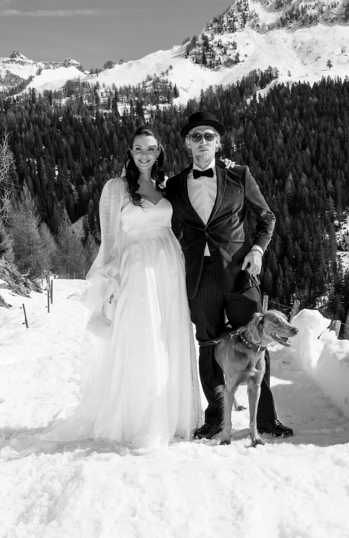 Ein Brautpaar in formeller Kleidung, begleitet von einem Hund, steht in einer verschneiten Berglandschaft.