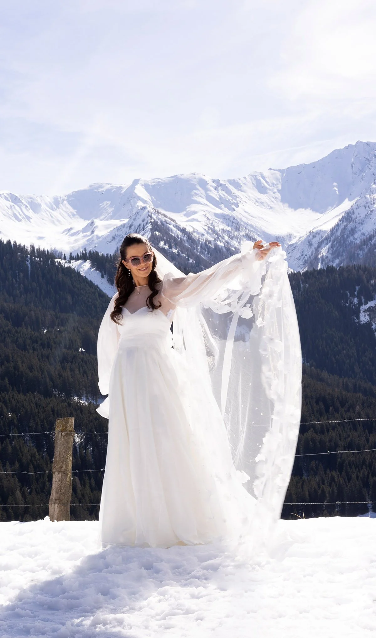 Eine Frau in einem weißen Hochzeitskleid steht im Schnee vor einer Bergkulisse mit schneebedeckten Bergen und Nadelbäumen, bei sonnigem Himmel, und hält ein weißes Tuch hoch.