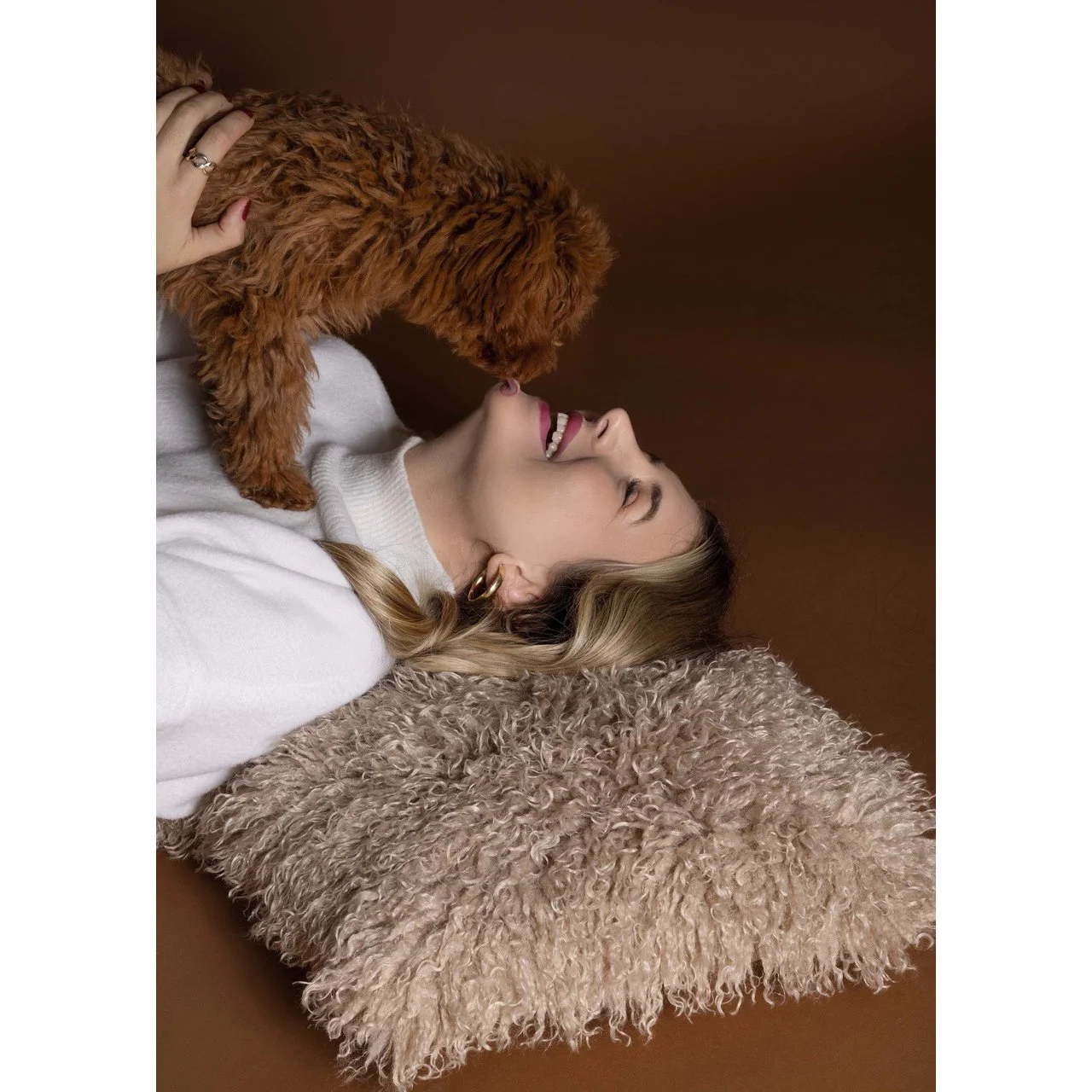 Eine lachende junge Frau liegt auf einem braunen Kissen, während ein brauner Hund ihren Kopf berührt.