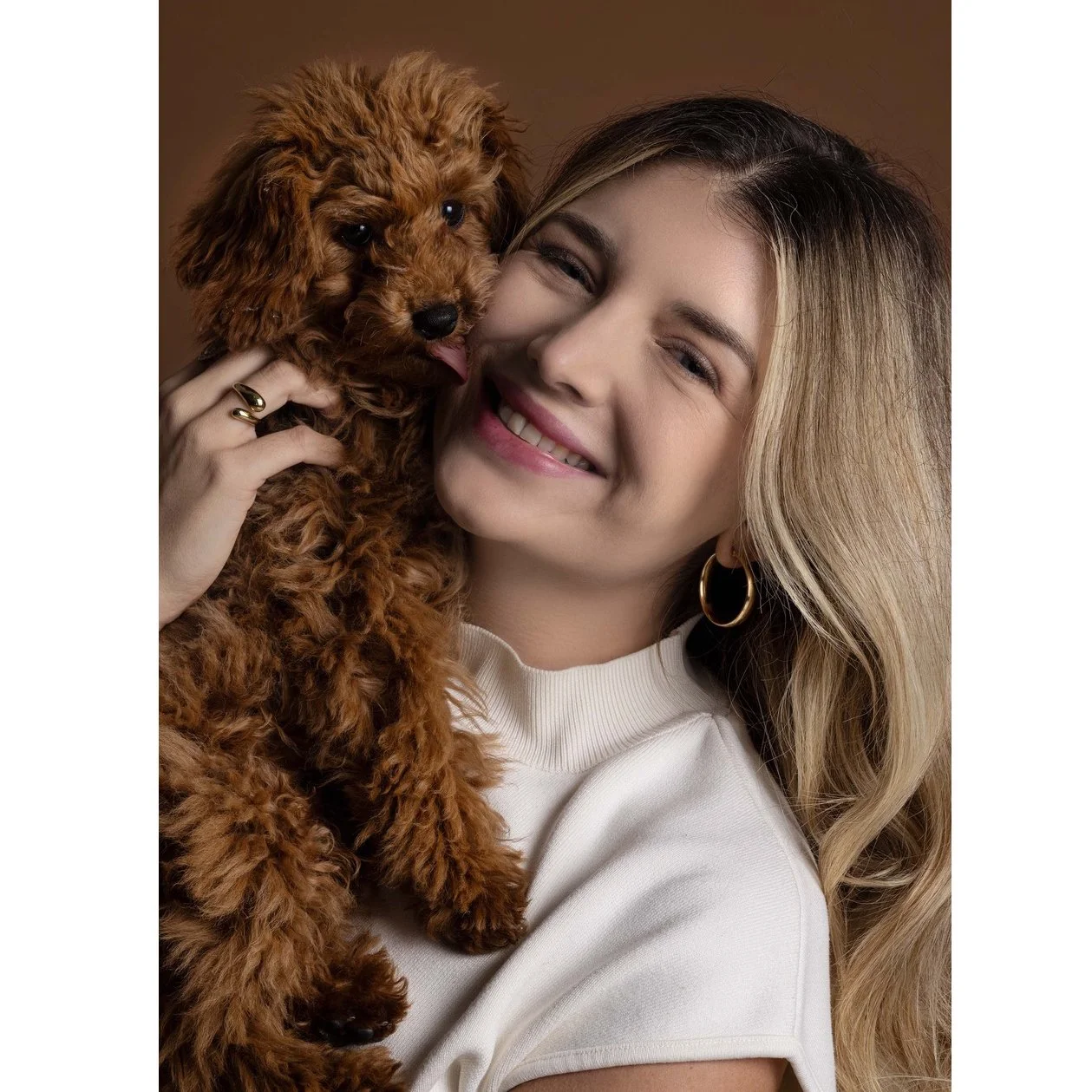 Lächelnde Frau hält einen kleinen braunen Hund in den Armen, beide schauen glücklich in die Kamera.